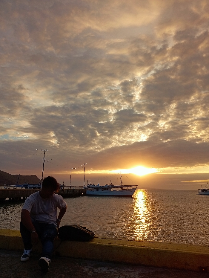 The image captures a sunset or sunrise over the harbor in Juan Griego, Venezuela. A male individual is seated in the foreground on a concrete pier, specifically on a yellow-painted curb or safety barrier. He wears a light-colored t-shirt, dark trousers, and athletic shoes. His head is slightly bowed, and he is looking towards the lower right of the frame. A dark bag rests on the pier beside his right side. The sky is largely covered with broken stratocumulus clouds, illuminated by a bright, low-hanging sun near the horizon, which casts a shimmering golden path across the calm dark water. Multiple dark poles with lighting fixtures extend along the pier to the left. In the midground, a light-colored boat with a mast and cabin is docked, reflecting the sunlight. Another vessel is partially visible further out on the water to the right, and smaller boats are discernible near the distant shoreline on the left, backed by a dark landmass.