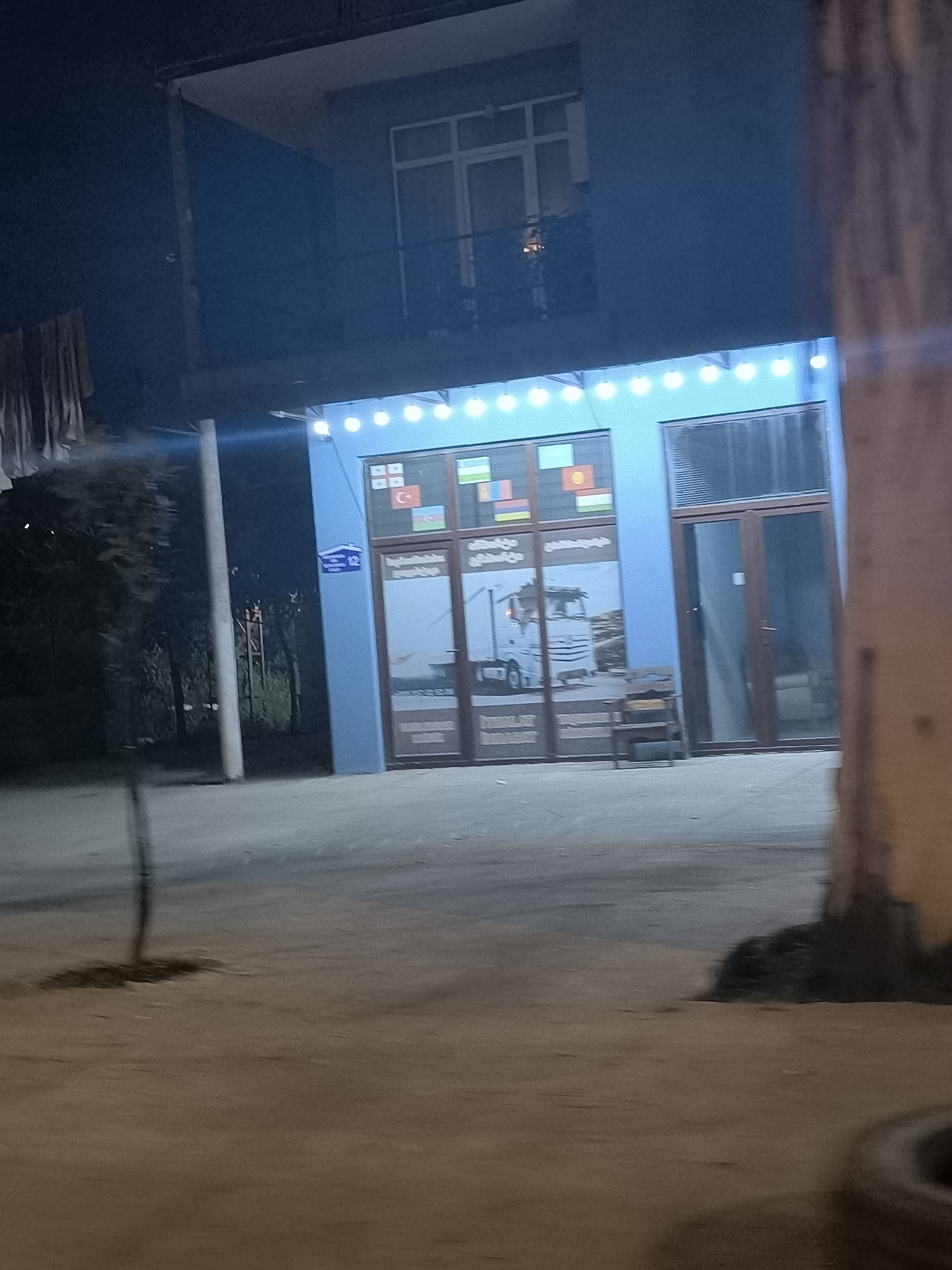 The image depicts a storefront in Batumi, Georgia, at night. The building has two stories, with the lower floor occupied by a business. The storefront features a large window displaying a truck and flags of several countries, including Georgia, Turkey, Uzbekistan, Armenia, and Kyrgyzstan. Above the window, a string of lights is illuminated. To the right of the storefront is a glass door. A street sign reading "12" is visible on the building. The scene is set on a paved surface, with trees and vegetation visible to the left. The overall impression is that of a commercial establishment in an urban setting.