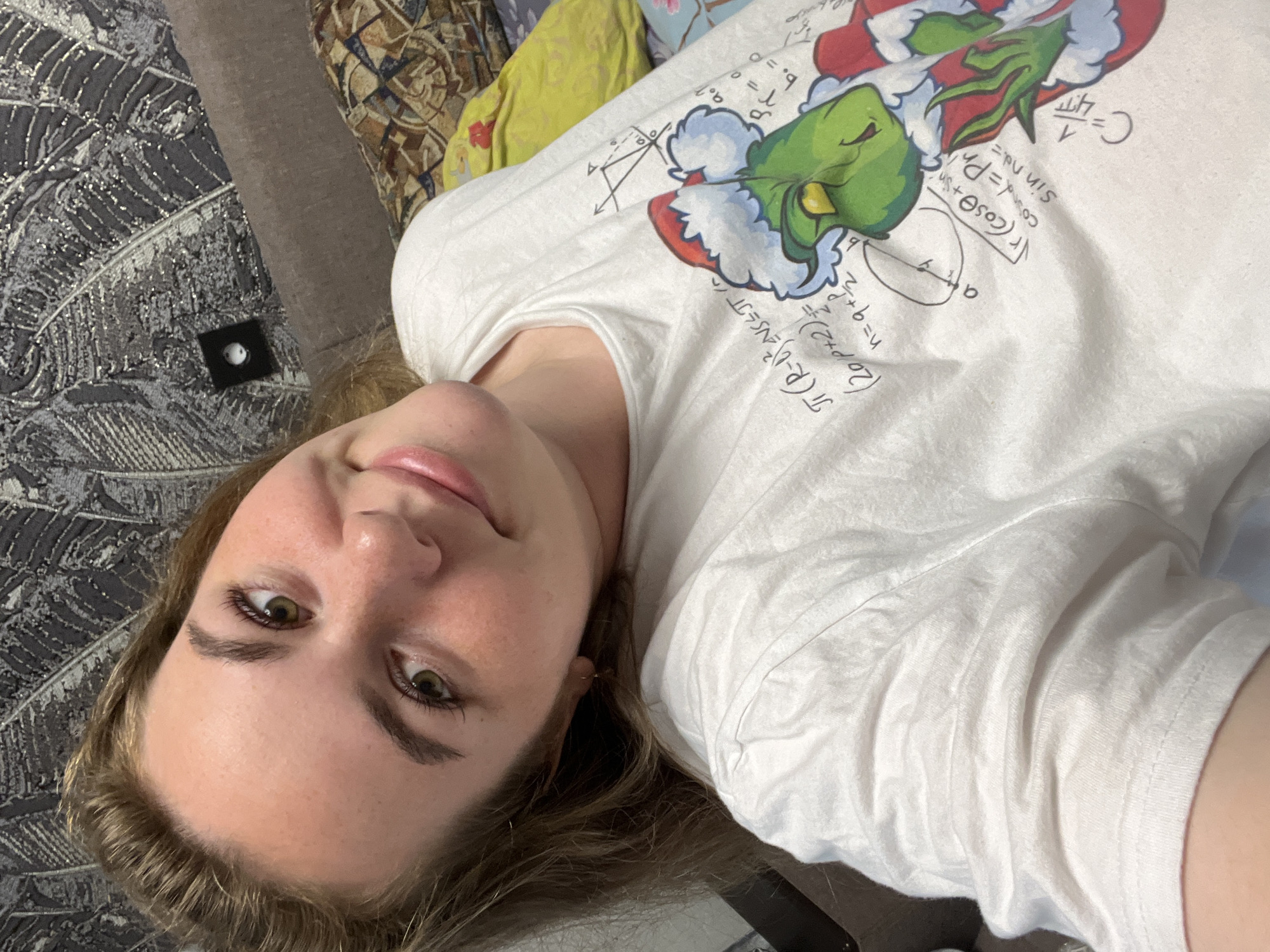 A young woman lies on a patterned grey and brown upholstered couch or bed. She wears a white t-shirt with a green and red Grinch graphic adorned with mathematical formulas and equations. The woman gazes directly at the camera with a slight smile. Her hair is blonde-brown. Behind her, there is a pillow with a yellow and floral print. The location is not identifiable as Chaikovsky, Russia.