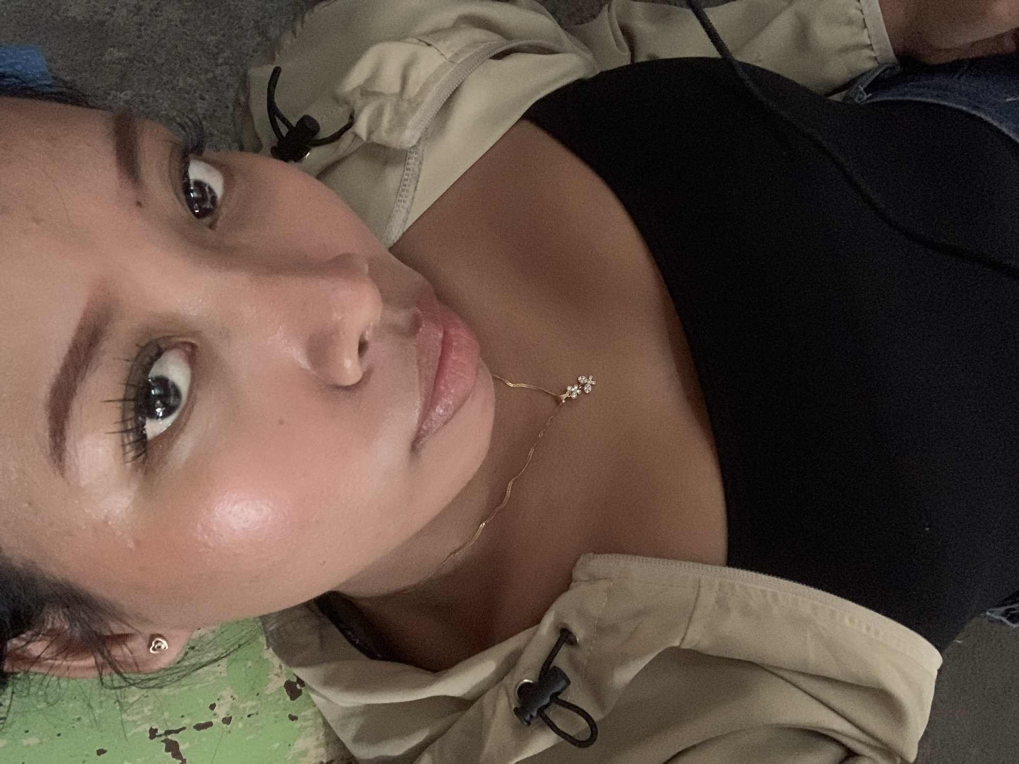 A close-up, upside-down, low-angle shot shows a young person lying down. They are wearing a black top and a light-colored jacket. A delicate gold necklace with two small flower charms is visible around their neck. The background consists of a gray surface, and to the right, a green, weathered surface with white speckles. The scene is not identifiable as being in Tamanbaru, Indonesia.