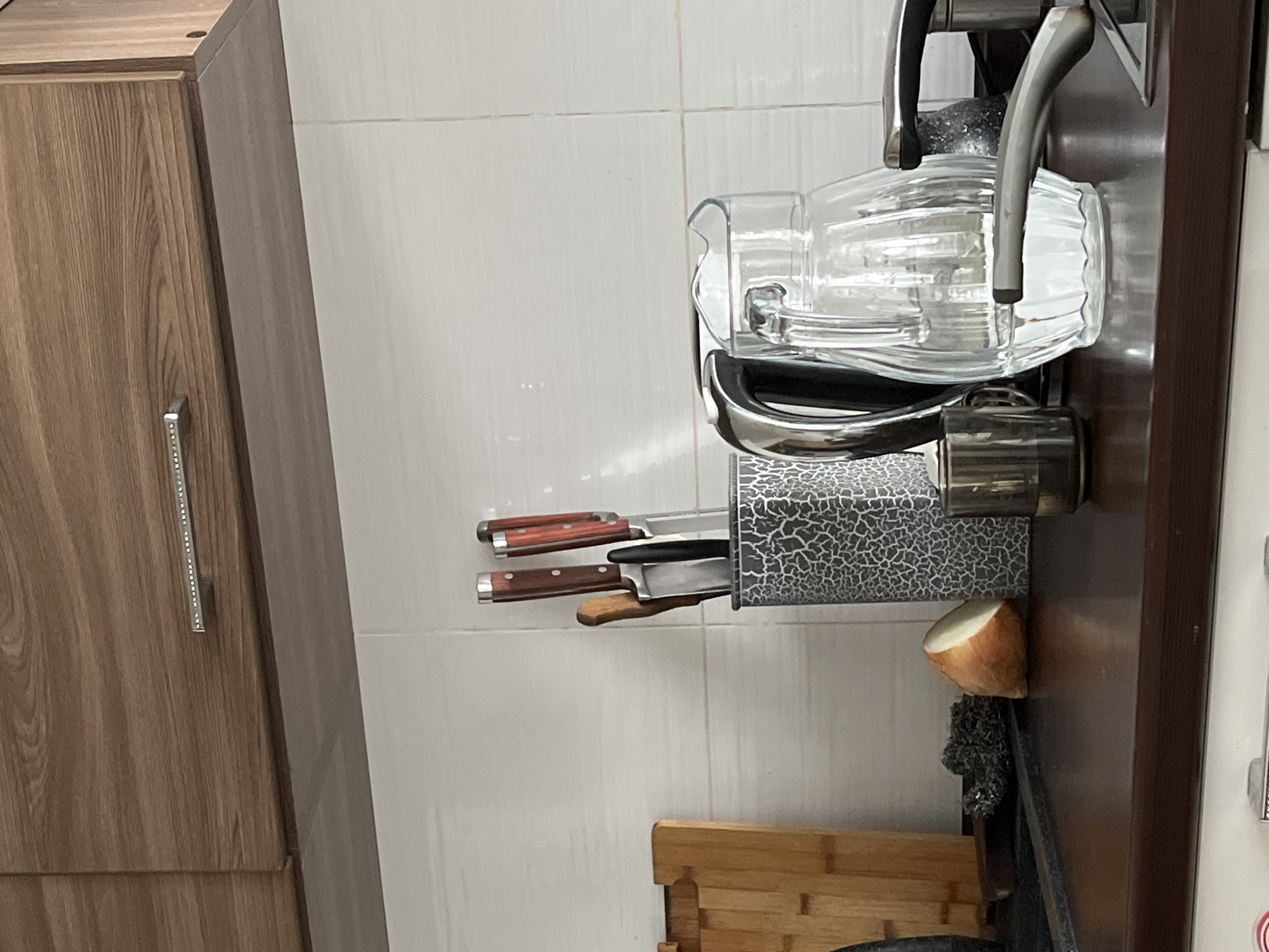 In an indoor kitchen setting, a dark brown countertop features a variety of items. To the left, a wooden cutting board with grooves is placed upright. Next to it, a metal scouring pad rests on the counter, followed by a cut onion with its white flesh exposed. A knife block, decorated with a crackled pattern, holds several knives with wooden handles. To the right of the knife block is a clear glass pitcher filled with water. Behind the pitcher, a metallic kettle with a black handle is visible. In front of the pitcher, a small, dark glass is present. The background consists of white tiled walls. Above the countertop, wooden cabinets with a horizontally grooved texture are mounted, one of which has a decorative silver handle with small embedded rhinestones.