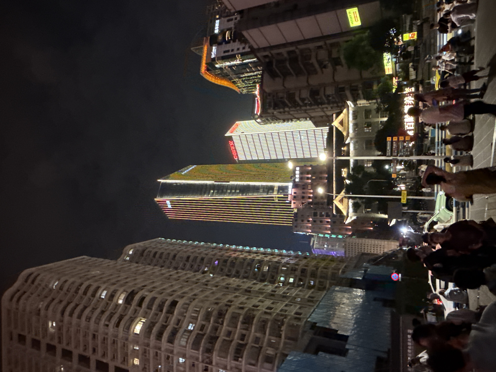 This is a nighttime, eye-level, wide shot of a street scene in the Yuzhong District of China. The foreground is populated with a crowd of people in casual clothing, some standing and some seated on a low barrier. A few cars are visible on the street behind them. Dominating the background are several tall buildings, some illuminated with colorful lights, and others appearing dark or partially lit. The sky is dark, indicating it is night. One building in the center features vertical yellow and pink lights. Another to its left has horizontal white lights. Further to the right, a building with an orange, curved roof is visible. The overall impression is of a bustling urban environment at night.