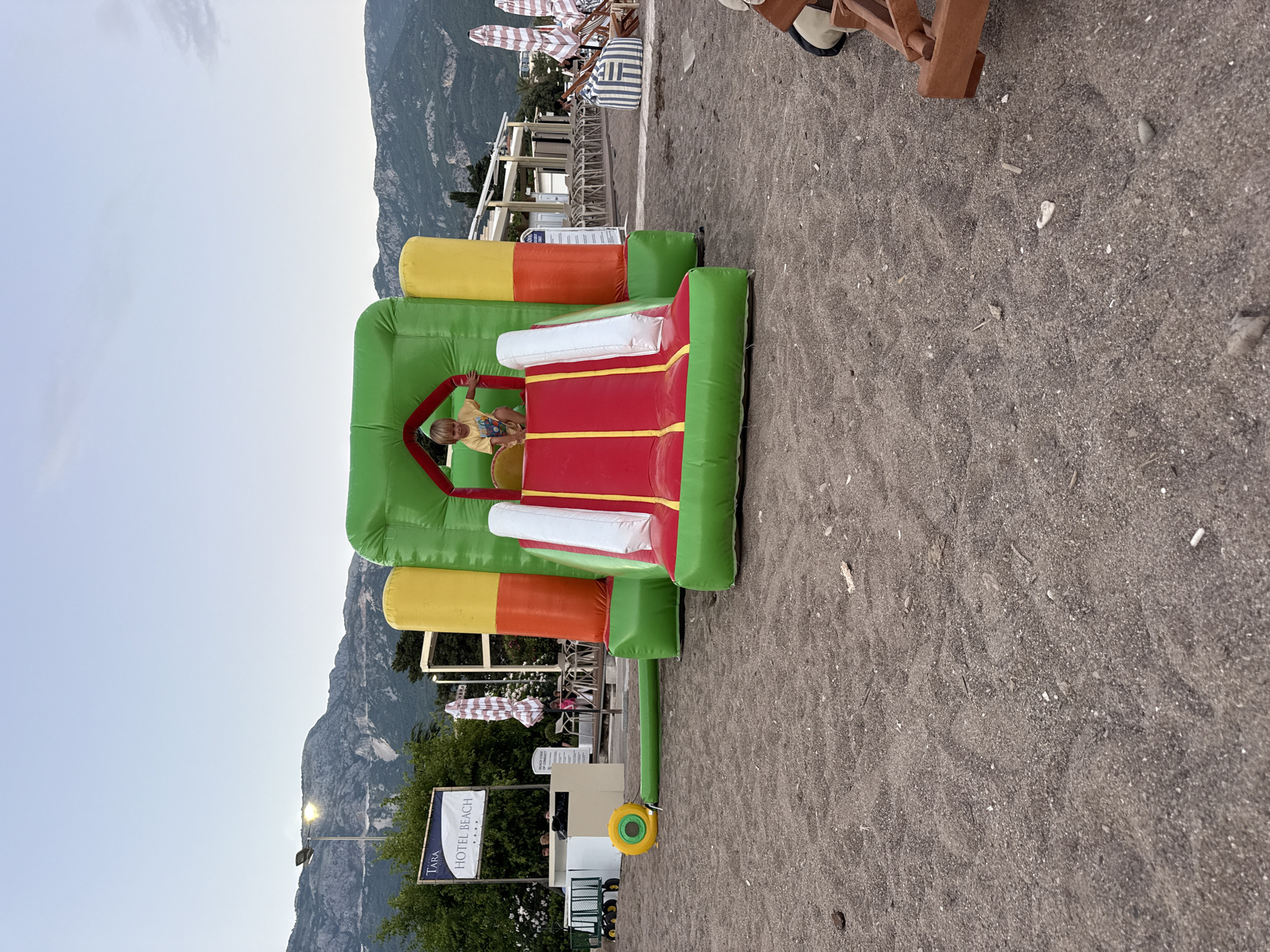 A young boy with blond hair, wearing a yellow t-shirt, is standing inside an inflatable bounce house on a sandy beach. The bounce house is predominantly green, with red and yellow sections, and has a slide. The beach is covered in dark sand, with some pebbles and debris scattered about. In the background, there is a sign for "TARA HOTEL BEACH", a lamppost, lush green trees, and a range of imposing mountains under a hazy sky. There are also beach chairs and umbrellas visible further in the background, suggesting a resort or hotel area. The scene appears to be set in Boreti, Montenegro.