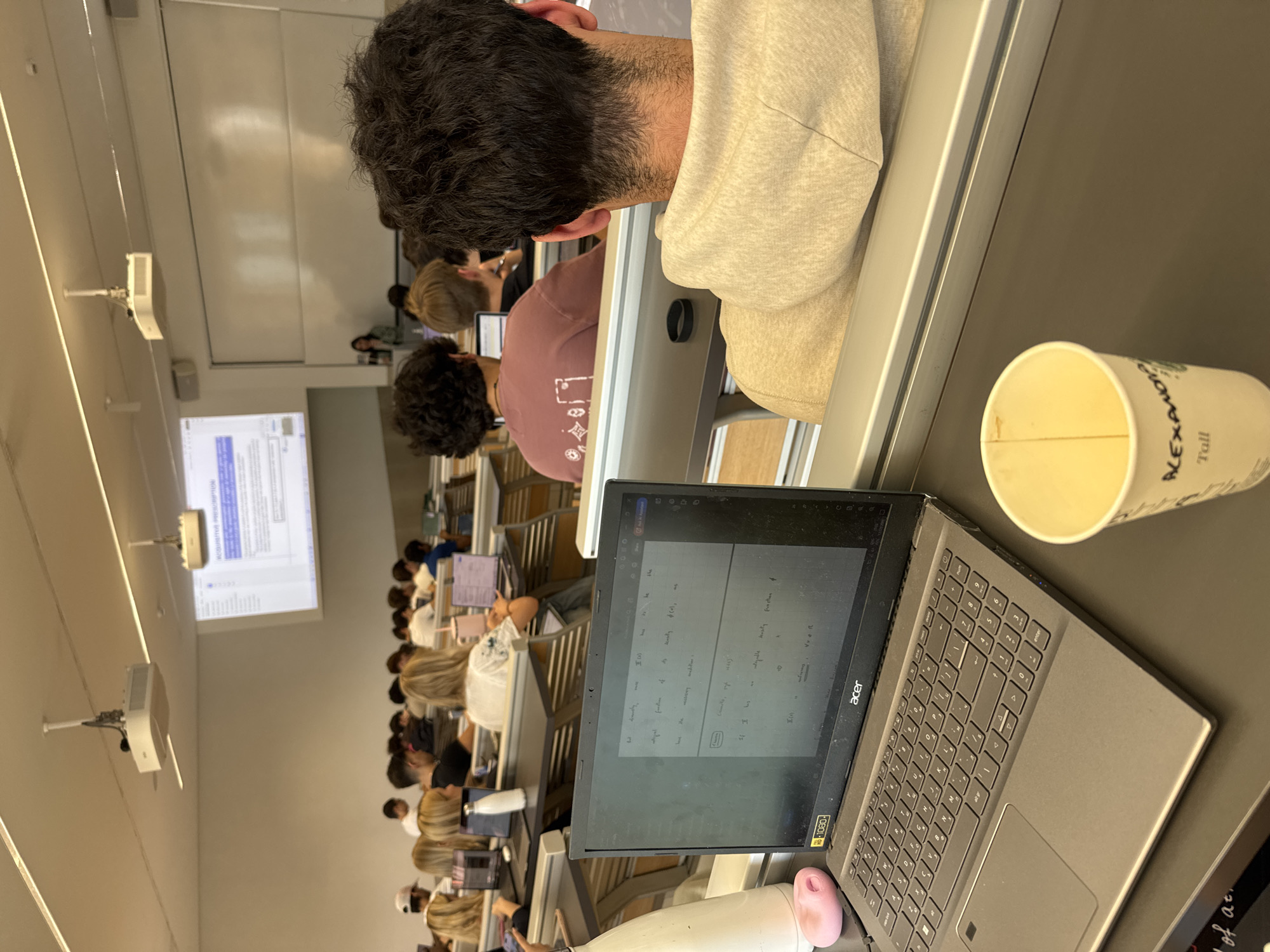 The image captures a classroom setting. Students are seated at desks equipped with laptops, suggesting a lecture or study session. A projection screen displays information at the front of the room. The presence of coffee cups implies a potentially long or early session. The environment appears to be a university lecture hall with fixed seating. There are no explicit indicators pointing to Milan, Italy, but the image depicts a typical university setting that could be located anywhere.