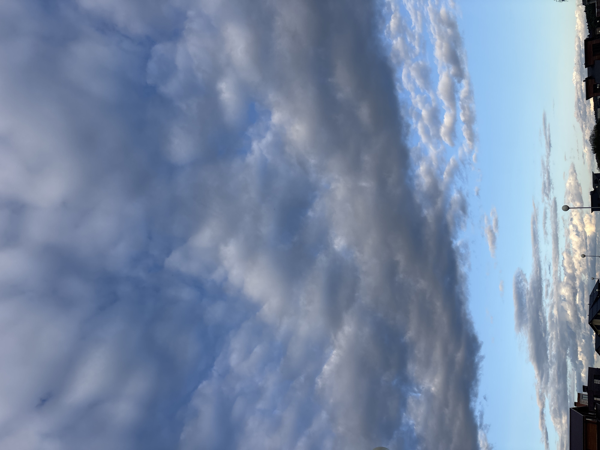 The image captures a view of the sky over the city of Zvenigorod, Russia, with a line of residential buildings along the lower edge. The sky is predominantly filled with a mix of clouds. Large, dark gray clouds dominate the upper portion of the frame, with some lighter blue sky peeking through in patches. Below these, there are smaller, scattered white and gray clouds. The horizon features the rooftops of several houses, characterized by dark-colored roofing materials. Streetlights are visible above the rooftops, one on the left and another on the right. The overall lighting suggests it is either early morning or late afternoon, with the sun not directly visible but casting light upon the clouds and structures. No people or actions are depicted in the scene.