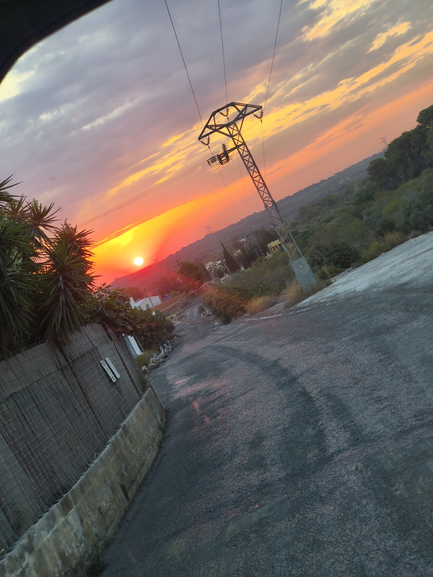 The image captures a sunset in Riba-roja de Túria, Spain. The sky is a vibrant mix of orange, yellow, and purple, with dramatic clouds. In the foreground, a paved road slopes downwards to the right. To the left, a wire mesh fence runs along a concrete wall, behind which are some palm-like plants and other vegetation. A power line tower stands prominently in the middle ground, with electrical wires stretching across the sky. Further in the distance, rolling hills covered in trees are visible, with a few scattered buildings and more power lines. The overall scene depicts a rural or semi-rural setting during sunset.