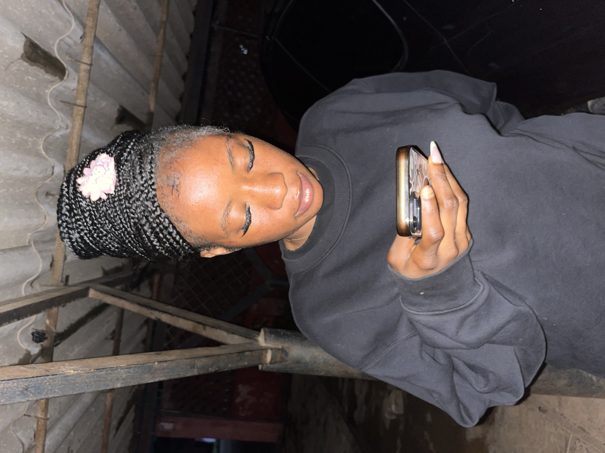 A young Black woman with braided hair and false eyelashes is looking down at a smartphone she is holding in her right hand. She is wearing a black sweatshirt. The background appears to be an outdoor, covered area with a corrugated metal roof supported by metal poles. The location is identified as Jos, Nigeria.