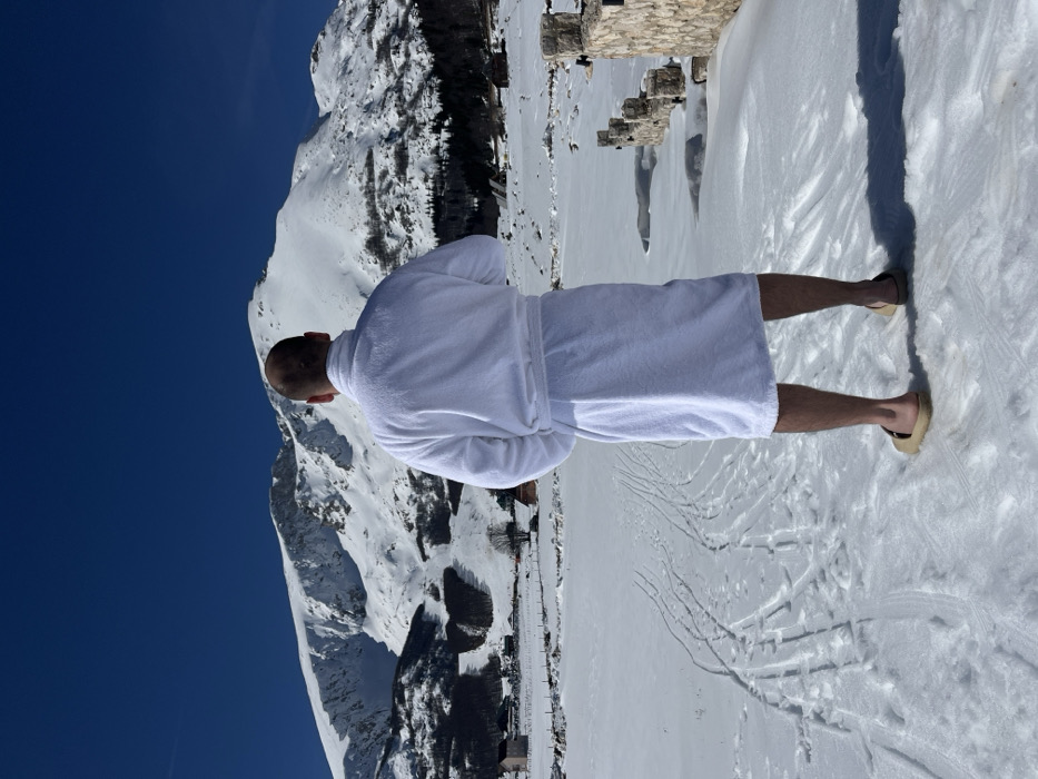 A male individual is depicted from behind, standing in an expansive snow-covered field under a clear blue sky in Virak, Montenegro. He is wearing a white terry cloth bathrobe, tied at the waist, with bare legs and light-colored open-toed sandals. His head exhibits short or shaved hair. The subject faces a panoramic view of large, snow-clad mountains in the background. On the lower slopes of the mountains, patches of evergreen trees, several small buildings, and visible ski slopes or cleared areas are present. The foreground is covered in snow, showing numerous tracks including footwear impressions and parallel lines consistent with skis or a sled, extending away from the camera. To the right of the subject, a low stone wall or structure with multiple square stone pillars is partially visible, protruding from the snow. A distinct shadow of the individual is cast onto the snow to his right.