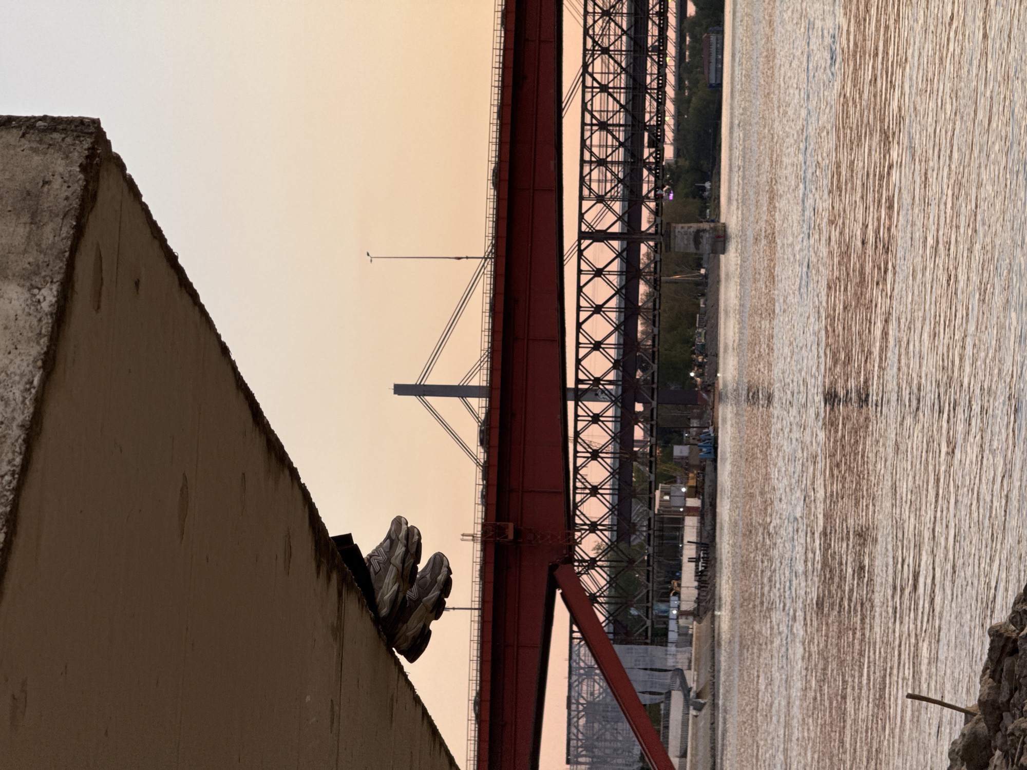 The image, when viewed in correct landscape orientation, captures a serene riverside scene bathed in the warm glow of either sunrise or sunset. On the left, a large, weathered concrete wall or structure occupies the foreground, its textured surface showing signs of age. Resting precariously on the uneven top edge of this concrete structure is a pair of grey sneakers, identified by the distinct "N" logo of the New Balance brand visible on the side.
Stretching across the midground is a large, imposing red metal bridge, characterized by its intricate truss design and sturdy support beams. A solitary street light can be seen on the bridge deck. Beneath and around the bridge on the distant bank, faint outlines of buildings, trees, and possibly construction or industrial elements are visible. To the right of the bridge, a wide body of water, likely a river, dominates the scene, reflecting the soft, warm light in shimmering, elongated streaks. The sky above is clear, displaying gentle hues of pale orange, pink, and yellow, indicative of the golden hour.
The scene appears peaceful and quiet, with no explicit human activity other than the presence of the abandoned sneakers, suggesting a moment of pause or contemplation. Given the location context, the bridge is likely one of the significant crossings over the Sava or Danube River in Belgrade, Serbia.