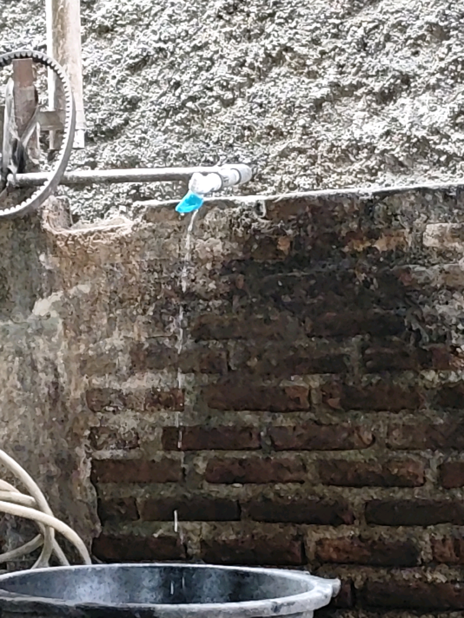 A close-up shot captures water dripping from a blue plastic nozzle attached to a white pipe. The pipe runs horizontally across the frame, positioned just below a rough, textured wall. Below the pipe and to the right, a weathered brick wall shows signs of age and moisture, with dark streaks marking where water has run down. In the lower left quadrant, a portion of a gray bucket and a coiled white hose are visible. To the left of the pipe, a metal gear-like object is partially in view, attached to a vertical post. The background consists of a coarse, greyish-white textured surface. The scene appears to be outdoors, possibly in a utility area, given the presence of plumbing and a bucket. There are no people or animals visible in the image. The location is in Kemuning, Indonesia.