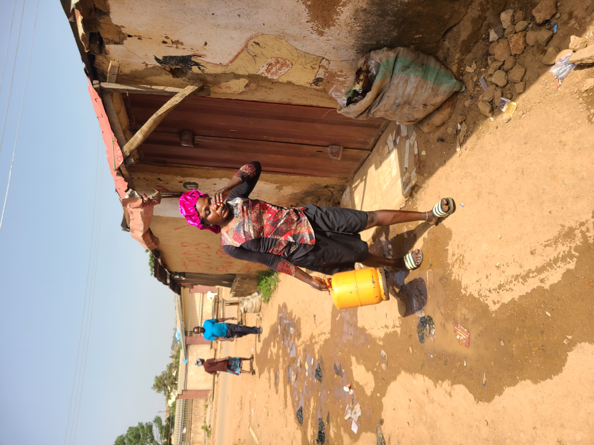 The image shows a woman standing in front of a building in what appears to be a poor neighborhood in Jos, Nigeria. She is wearing a pink headscarf, a long-sleeved red patterned shirt, and black shorts. She is holding a yellow gas tank in her left hand and touching her forehead with her right hand. The ground around her is wet and littered with trash. In the background, two people are walking down the street. The building behind her is dilapidated, with a rusty red roof and peeling paint. A large bag is propped up against the wall next to the door.