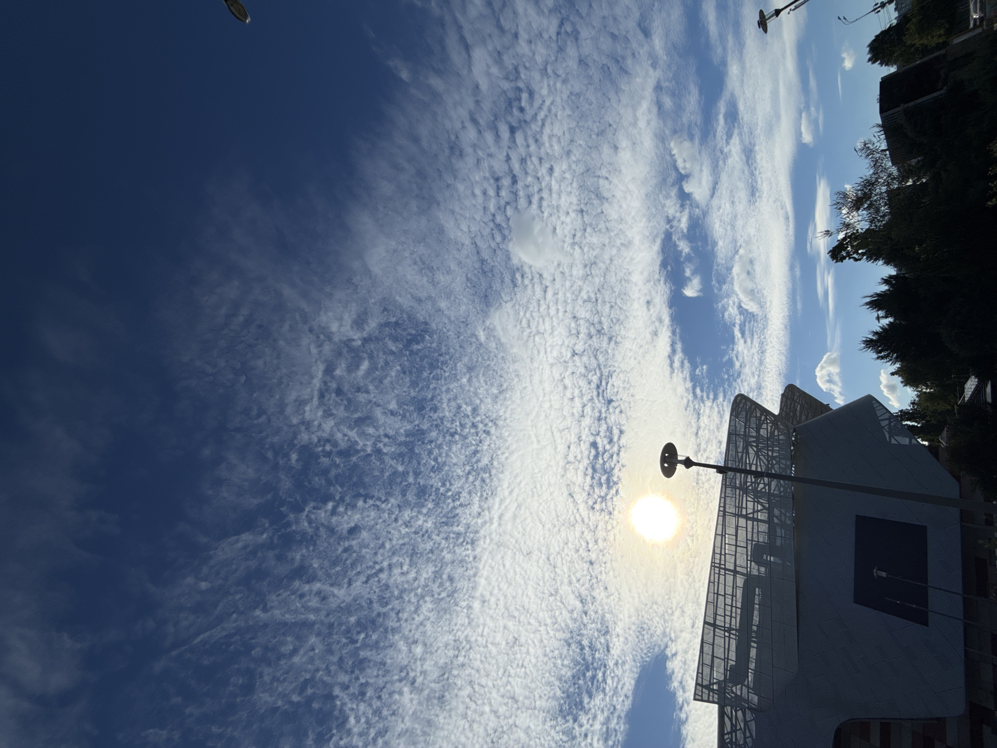 The image captures a bright, sunny day with a vast, blue sky filled with scattered, wispy white clouds. The sun is prominently visible, casting a bright glare. In the foreground, the angular, modern architecture of a building, possibly a stadium or cultural center, dominates the lower left portion of the frame. The building's facade appears to be made of metal and glass. A tall lamppost stands in front of the building, its light fixture silhouetted against the sky. Further to the right, the tops of dark green trees are visible, suggesting an urban park or green space. The scene appears to be in Moscow, Russia, given the architectural style and the general atmosphere. There are no people visible in the image.