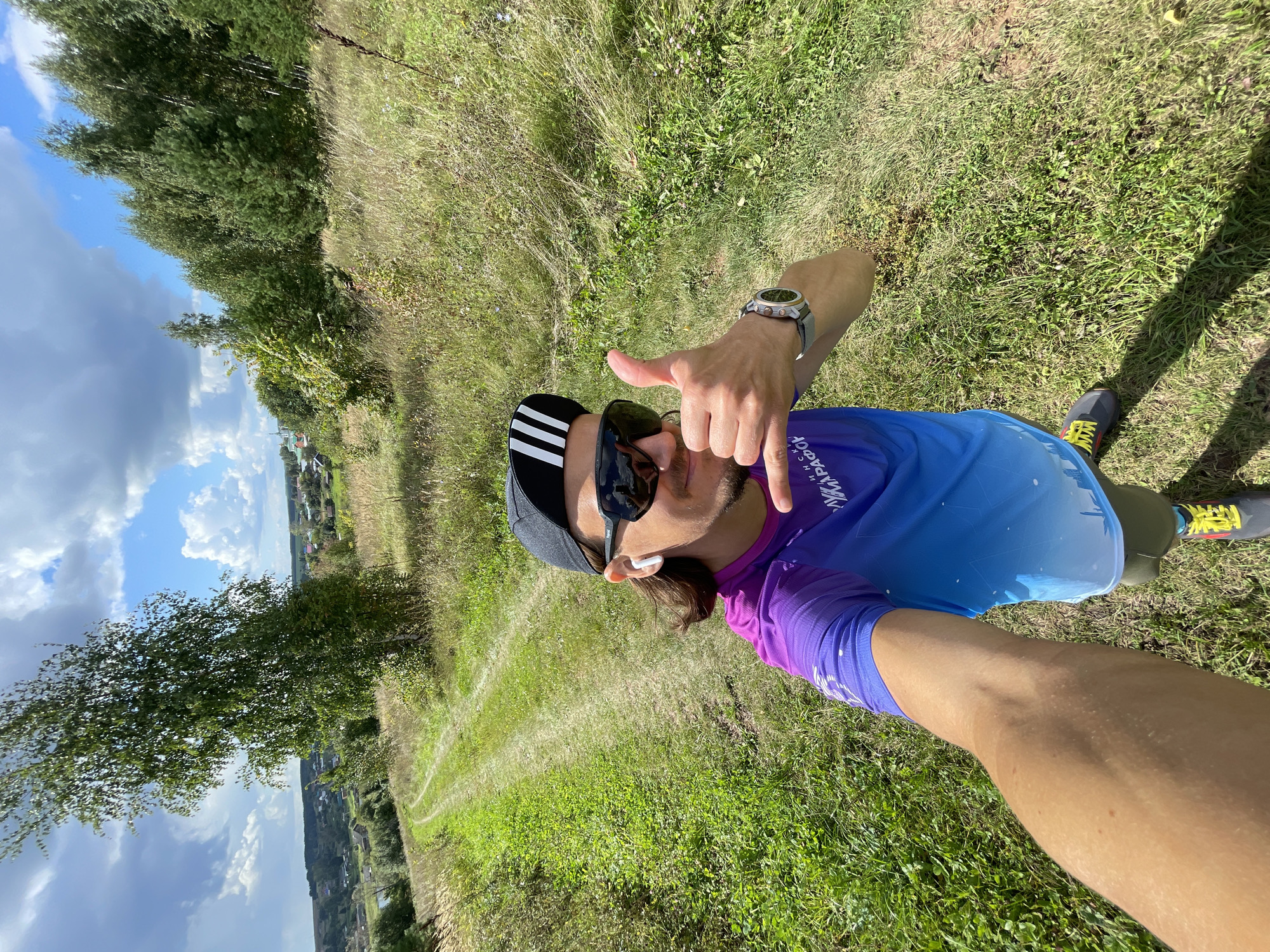A selfie taken by a person standing on a grassy dirt road in a rural area. The person is wearing a black baseball cap with white stripes, sunglasses, and a colorful athletic shirt. They are making a hand gesture that resembles the "shaka" sign and giving a thumbs-up. A smartwatch is visible on their left wrist. The background features a wooded area with trees, fields, and a distant view of a village. The sky is partly cloudy. The location appears to be in or near Perevoznoe, Russia, based on the provided hint.