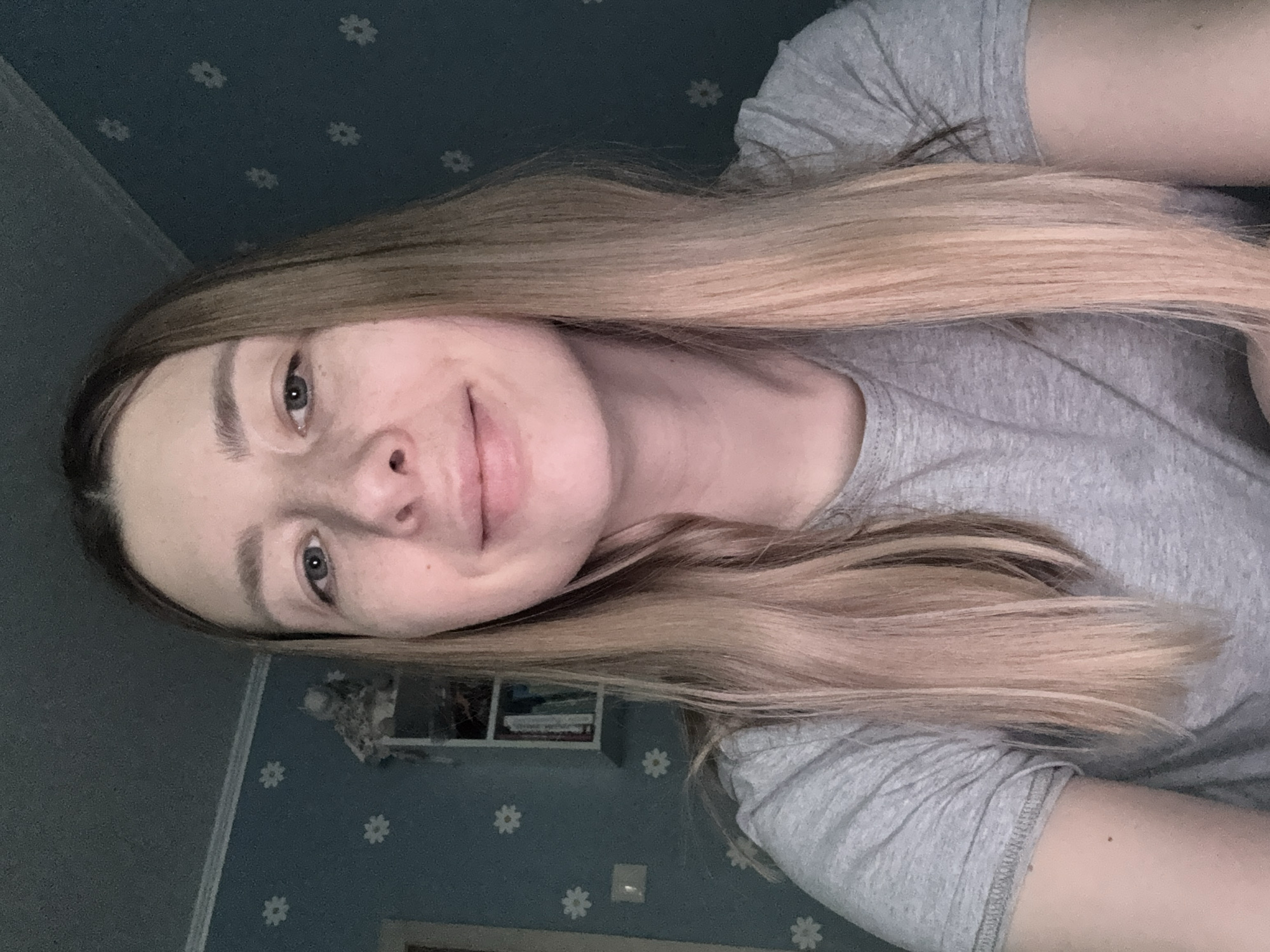 A fair-skinned woman with light brown hair is shown from the chest up. She is wearing a gray t-shirt. The background consists of a blue wall with white floral patterns. A shelf with books and decorative objects is visible behind her on the wall. The location cannot be determined from the image.