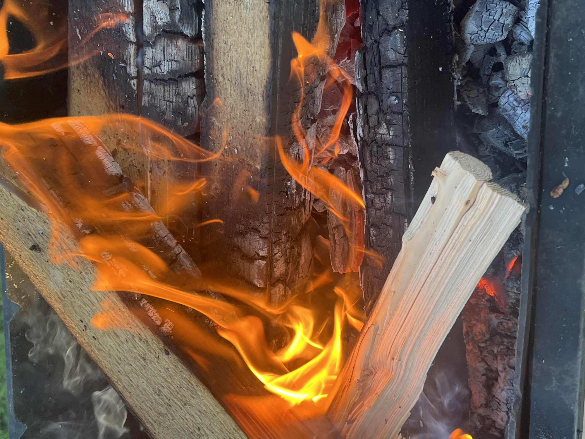 The image shows a close-up view of a fire burning in a metal fire pit. The fire is intense with bright orange and yellow flames licking at the logs. The logs themselves are a mix of freshly cut wood and charred remnants, indicating that the fire is actively burning and consuming the fuel. The background is dark and out of focus, suggesting the photograph was taken in low light conditions, possibly at dusk or night. The overall impression is one of raw heat and power. There is no visible indication of the city of Il'ino, Russia, in this image.