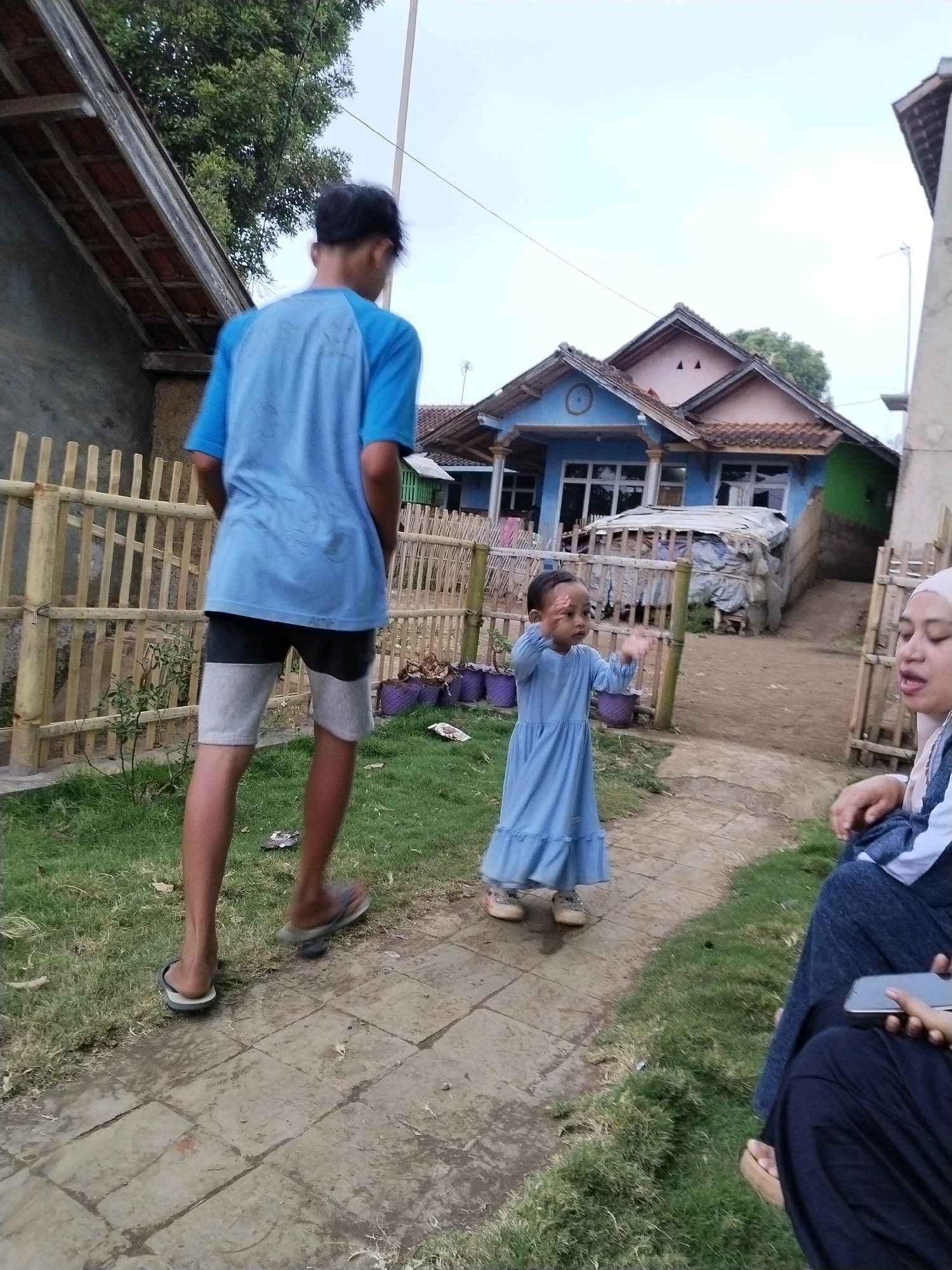 A group of people, likely Indonesian, are gathered outdoors in what appears to be a residential area in Garut, Indonesia. In the foreground, a teenage boy in a blue t-shirt and grey and black shorts stands facing away from the camera, looking towards a small child. The child, wearing a light blue dress, is facing the boy with their arms outstretched. To the right of the child, a woman wearing a headscarf and blue and white clothing is seated, looking to her right and holding a smartphone. The background features a light blue house with a pink trim, a green extension, and a brown tiled roof. A bamboo fence runs along the left side of the image, separating the grassy yard from the paved walkway. Several purple planters and some debris are visible near the fence. The scene is illuminated by natural light, suggesting it is daytime.