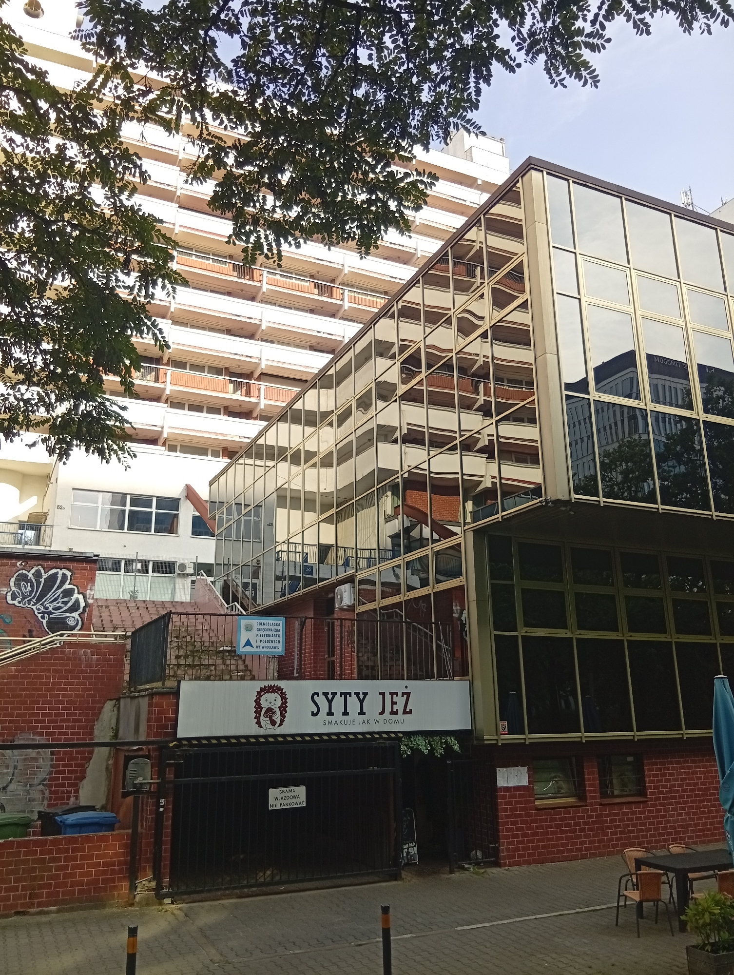 The image shows the facade of a modern building with large, reflective glass windows. The building appears to be a commercial establishment, with a sign that reads "SYTY JEŻ SMAKUJE JAK W DOMU" (which translates to "The Satisfied Hedgehog Tastes Like Home"). The entrance to the establishment is visible, with a metal gate and an intercom system.
In the background, a taller, older building with balconies and rows of windows is visible. Trees with green leaves partially obscure the view of the buildings, casting shadows on the scene. The sky above is light blue with some wispy clouds.
A sign near the entrance indicates "DOLNOŚLĄSKA OKRĘGOWA IZBA PIELĘGNIAREK I POŁOŻNYCH WE WROCŁAWIU" (Lower Silesian District Chamber of Nurses and Midwives in Wrocław). This confirms the location as Wrocław, Poland. The overall scene depicts an urban environment with a mix of modern and older architecture. There are no people visible in the immediate foreground, though the presence of outdoor seating with chairs and tables suggests a public space.