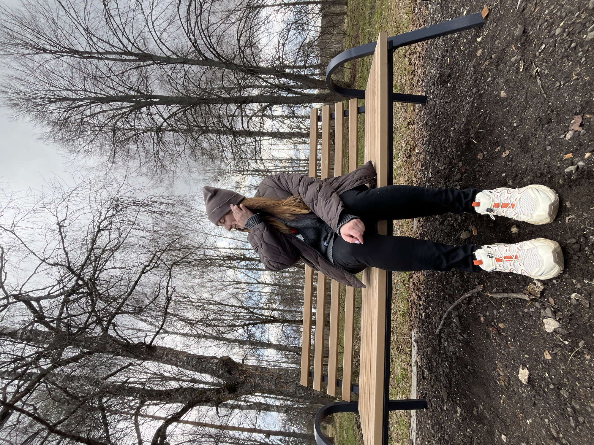 A woman in a brown hat and jacket sits on a wooden bench in a park. She is wearing black pants and white sneakers with orange accents. The ground around the bench is dirt and pebbles. Bare trees fill the background, and the sky is overcast. The location is Vysokovsk, Russia.