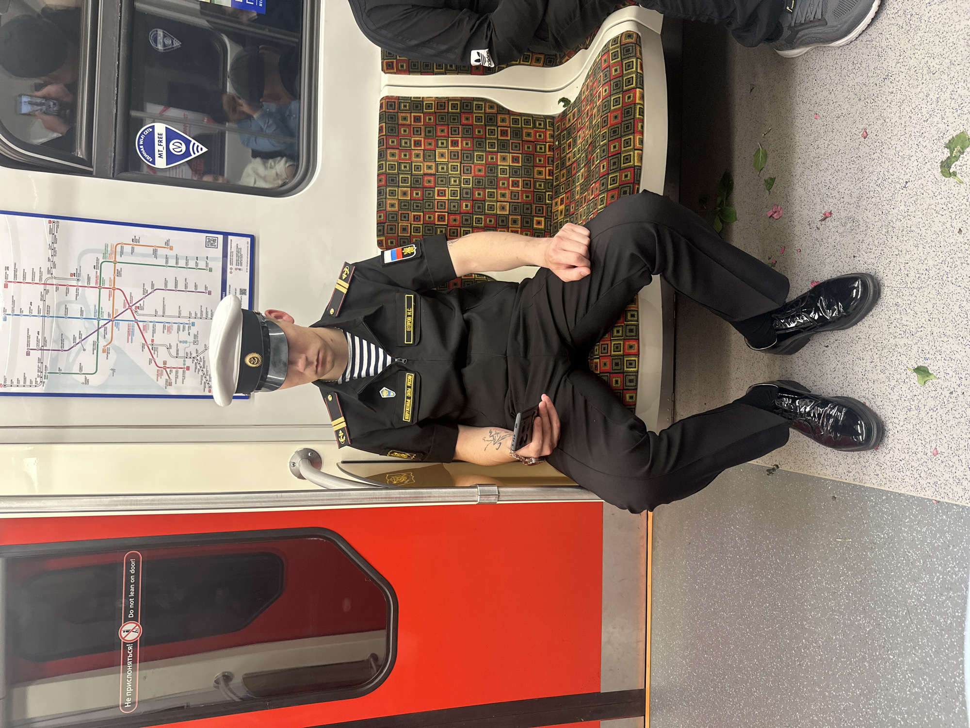 A man in a Russian naval uniform sits on a subway car seat in Saint Petersburg, Russia, holding a smartphone. The man is wearing a white naval cap, a dark uniform jacket with striped collar insignia and epaulets, and dark trousers. He is seated on a patterned subway seat with geometric designs in red, yellow, and black. To his left is a red subway door with a warning sign in Russian and English stating "Do not lean on door!". In the background, a subway map is visible on the wall. The floor of the subway car is gray and speckled. There are some fallen pink petals and green leaves on the floor.