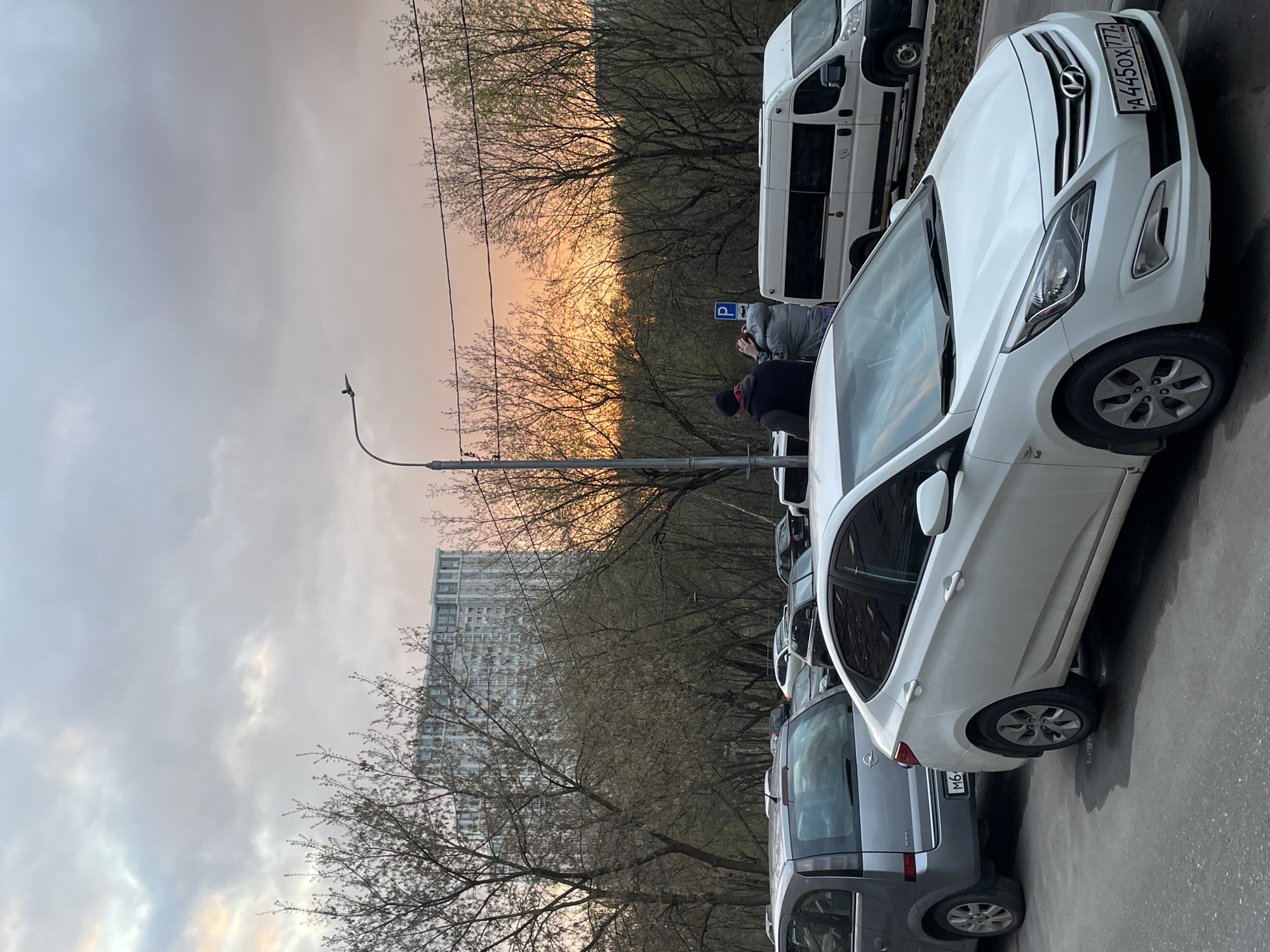 This image, captured in Moscow, Russia, is oriented sideways, appearing as if rotated 90 degrees clockwise. It depicts a street scene during either sunrise or sunset, characterized by a striking sky that transitions from soft grey and blue to a vibrant orange and pink glow near the horizon. This warm light backlights the numerous bare trees lining the street, some showing early signs of new foliage.
In the foreground, a white Hyundai Accent is prominently parked, with its Russian license plate "A445OX 777 RUS" clearly visible on the front. Behind the Hyundai, a white commercial van is parked, and a grey Opel Meriva is partially visible further down the line of cars. Two individuals are standing near the white van; one wears a dark jacket and a hat, while the other is dressed in a lighter grey jacket. A blue square parking sign with a white "P" symbol is visible next to the van.
A tall street lamp post extends into the frame from the middle-left. In the background, beyond the leafless trees, a large, multi-story building with numerous windows stands, suggesting an urban residential or office environment. The scene is static, portraying a quiet moment during twilight.