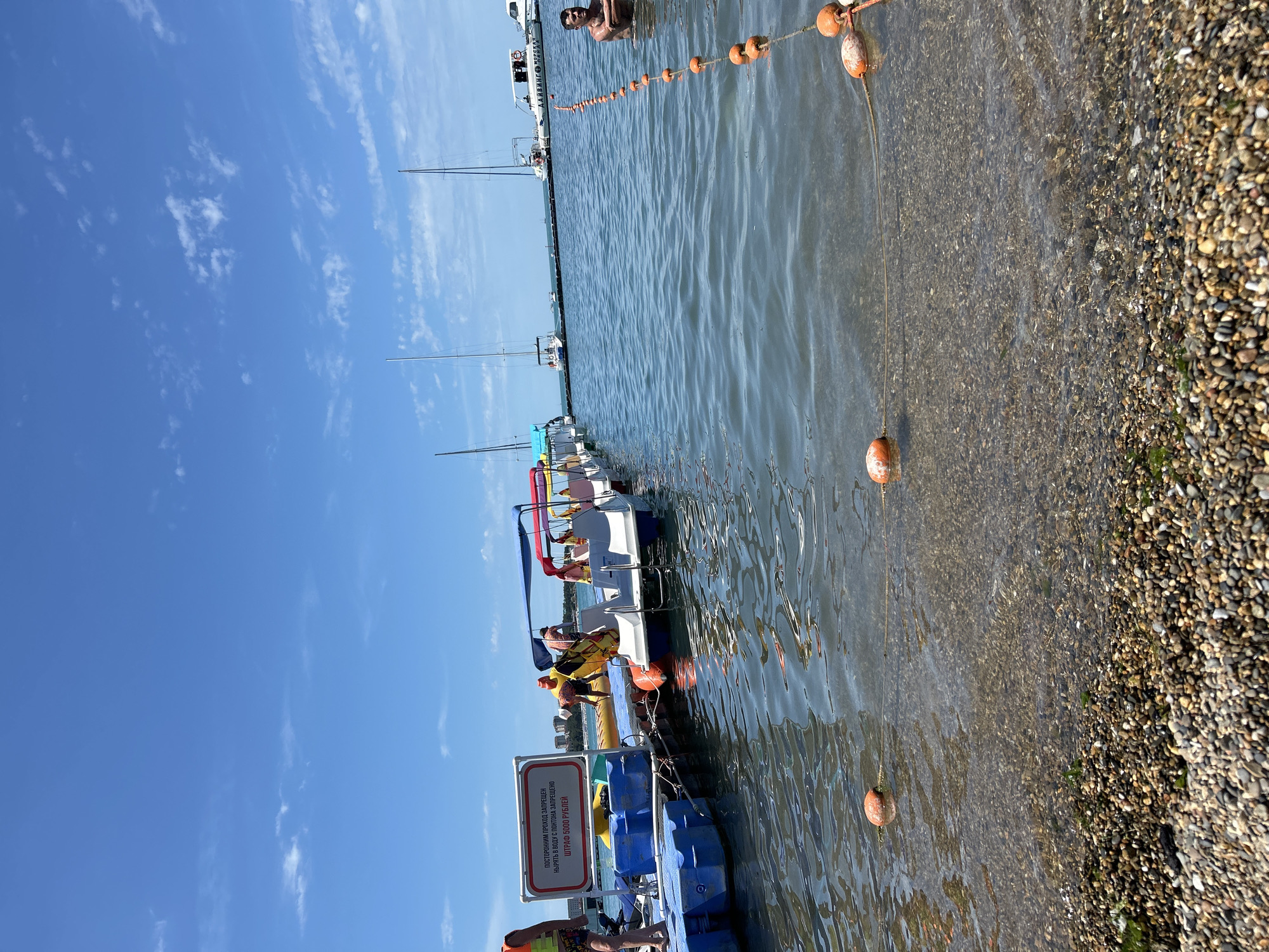 The image depicts a sunny day at a waterfront location, likely a beach or harbor, in Gelendzhik, Russia. The scene features a pontoon dock with several small watercraft, possibly pedal boats or small motorboats, lined up. People are visible on the dock and in the water. One person wearing a life vest stands on the pontoon. Several individuals are seated in or near the boats. In the background, sailboats and a larger boat with text that appears to read "ДАРВИН МОРСКАЯ" are anchored. A rope with orange buoys extends into the water, marking a boundary or swimming area. The water is clear, revealing a pebbly seabed near the shore. A sign on the pontoon reads "ПОСТОЯННЫМ ПРОХОД ЗАПРЕЩЕН" (Permanent passage is prohibited) and "НЫРЯТЬ В ВОДУ С ПЛОТИНА ЗАПРЕЩЕНО" (Diving into the water from the dams is prohibited) with a penalty of "ШТРАФ 6000 РУБЛЕЙ" (Fine 6000 rubles). The sky is blue with scattered white clouds. The overall impression is of a recreational area by the sea under a clear sky.