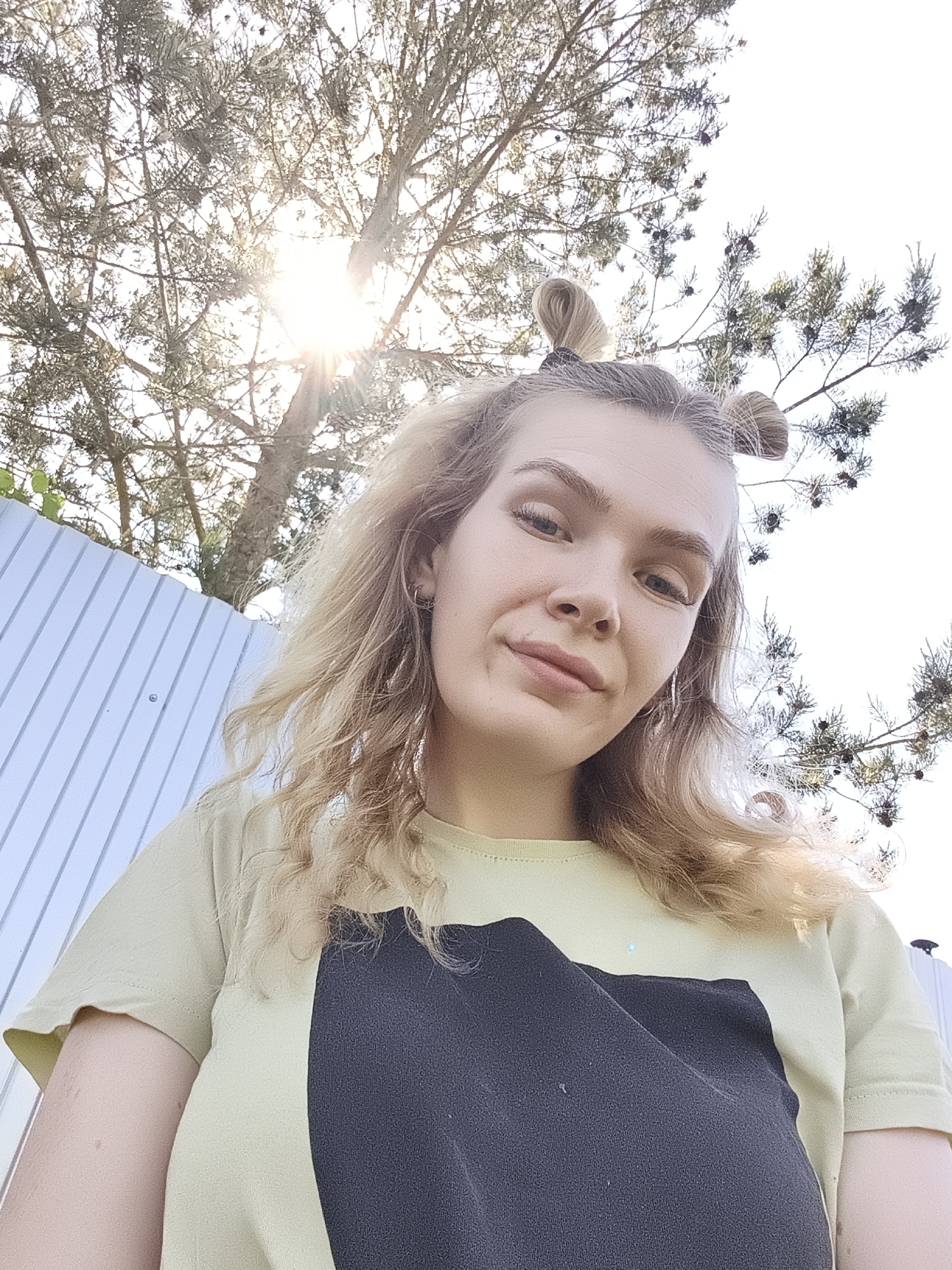 A low-angle shot captures a person with blonde hair styled in two small buns atop their head. They are wearing a light green t-shirt with a large black rectangle on the front. The person is positioned outdoors with a view of a tree extending overhead. Sunlight filters through the branches, creating a bright glow in the upper part of the frame. In the background, a white corrugated metal fence is partially visible. The location is Русский Вожой, Russia.