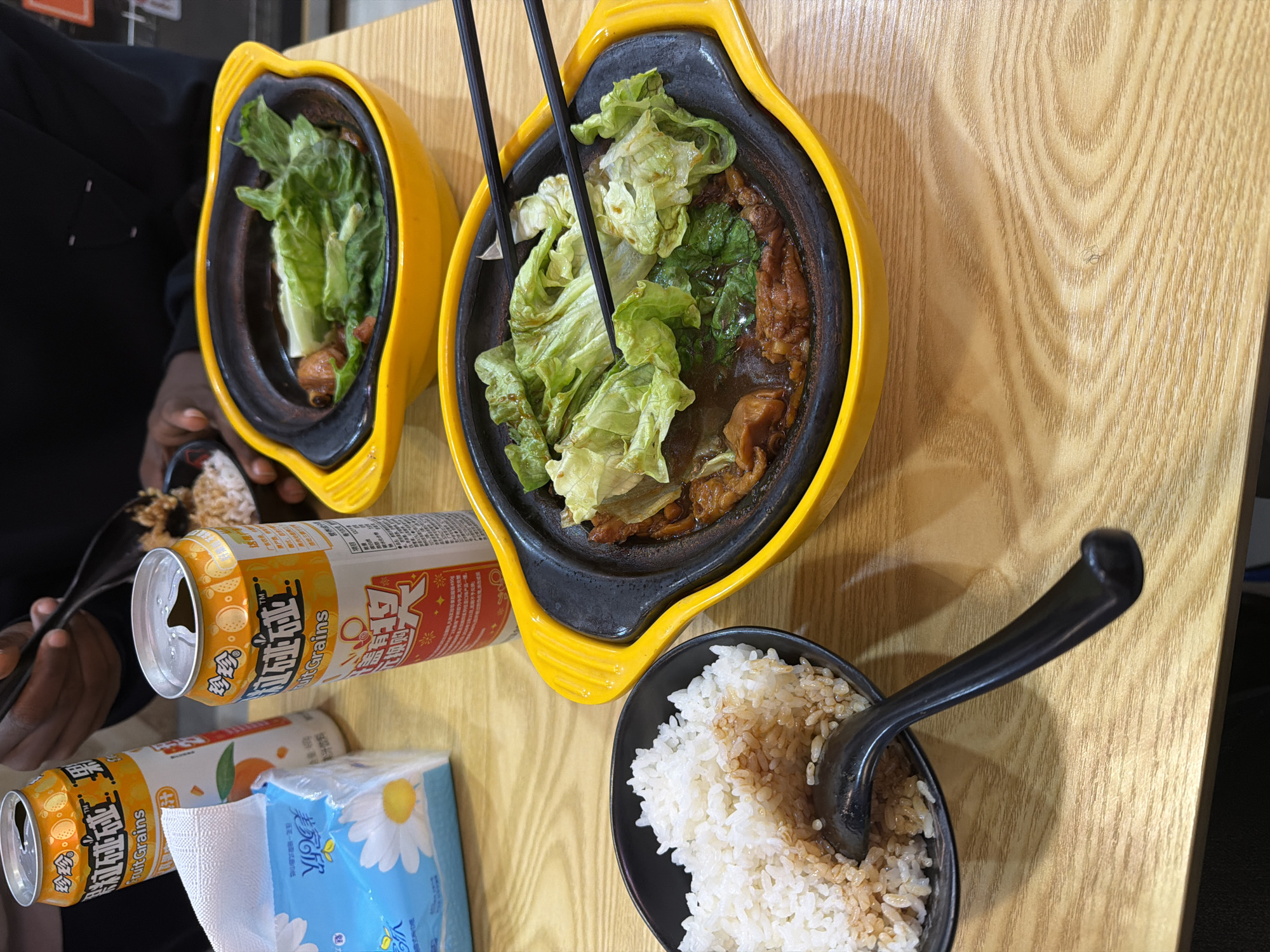 Here is a detailed description of the image:
Scene: An overhead shot of a table in Jiangning District, China.
Subjects and People: The dark-skinned hands of a person are visible at the left of the frame, holding a black bowl containing rice mixed with brown sauce.
Objects:
- Two oval-shaped bowls, each containing what appears to be a mix of cooked food and lettuce. One bowl has chopsticks sticking out of it. These bowls have a yellow exterior and a dark brown interior.
- A small black bowl of rice with a black spoon resting in it.
- Two cans of "Fruit Grains" beverage. One can is open.
- A package of tissues with a blue and white daisy design.
Environment:
The table is made of light-colored wood. The overall setting is a dining environment.