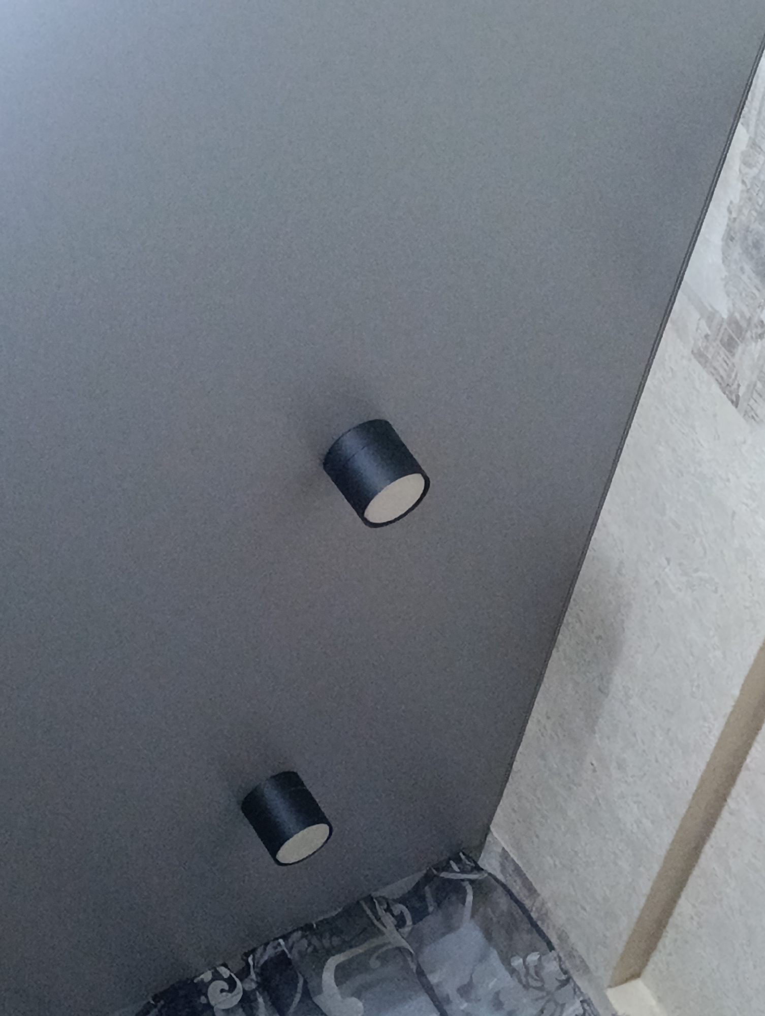 The image shows a gray ceiling with two cylindrical black spotlights. The ceiling meets a textured white wall on the right side. Below the ceiling, there is a glimpse of patterned bedding. There is no indication of the location being Krasnoyarsk, Russia, or any other location. There are no people, actions, or interactions visible in the image.
