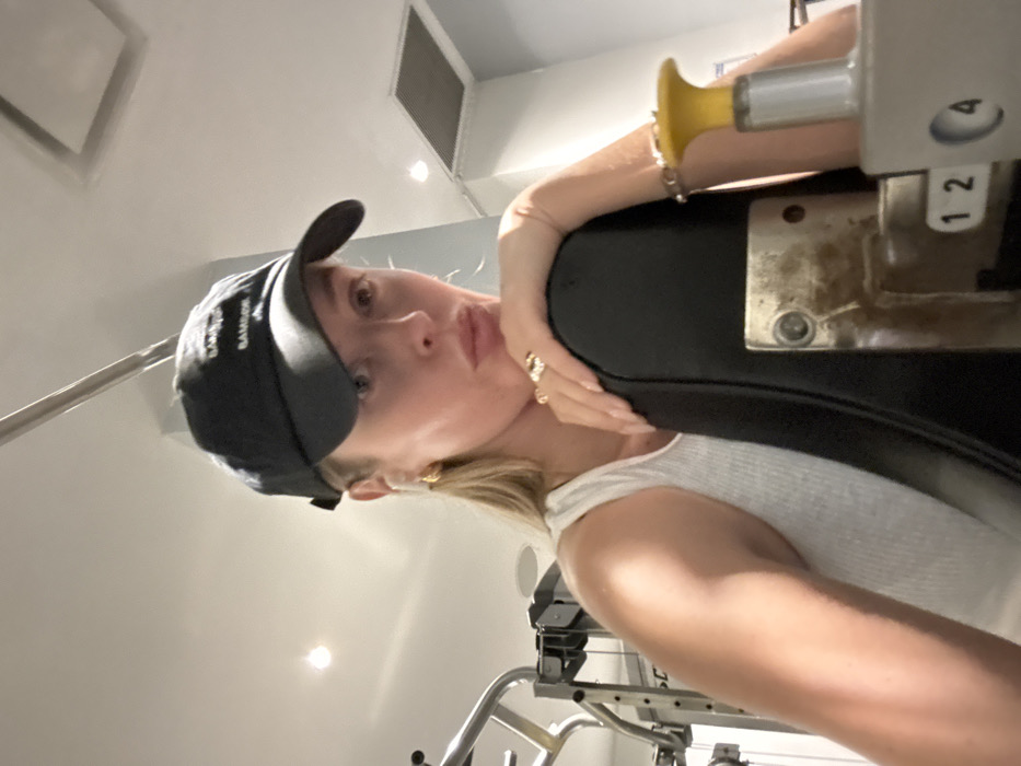 A woman with blonde hair wearing a black baseball cap and a white tank top poses for a selfie in a gym. She is leaning against a piece of gym equipment that has a black pad and a silver adjustment mechanism with a yellow handle. Her left arm is resting on the pad. A bracelet and rings are visible on her left hand. The background shows a bright, minimalist gym setting with white walls and rectangular ceiling panels.