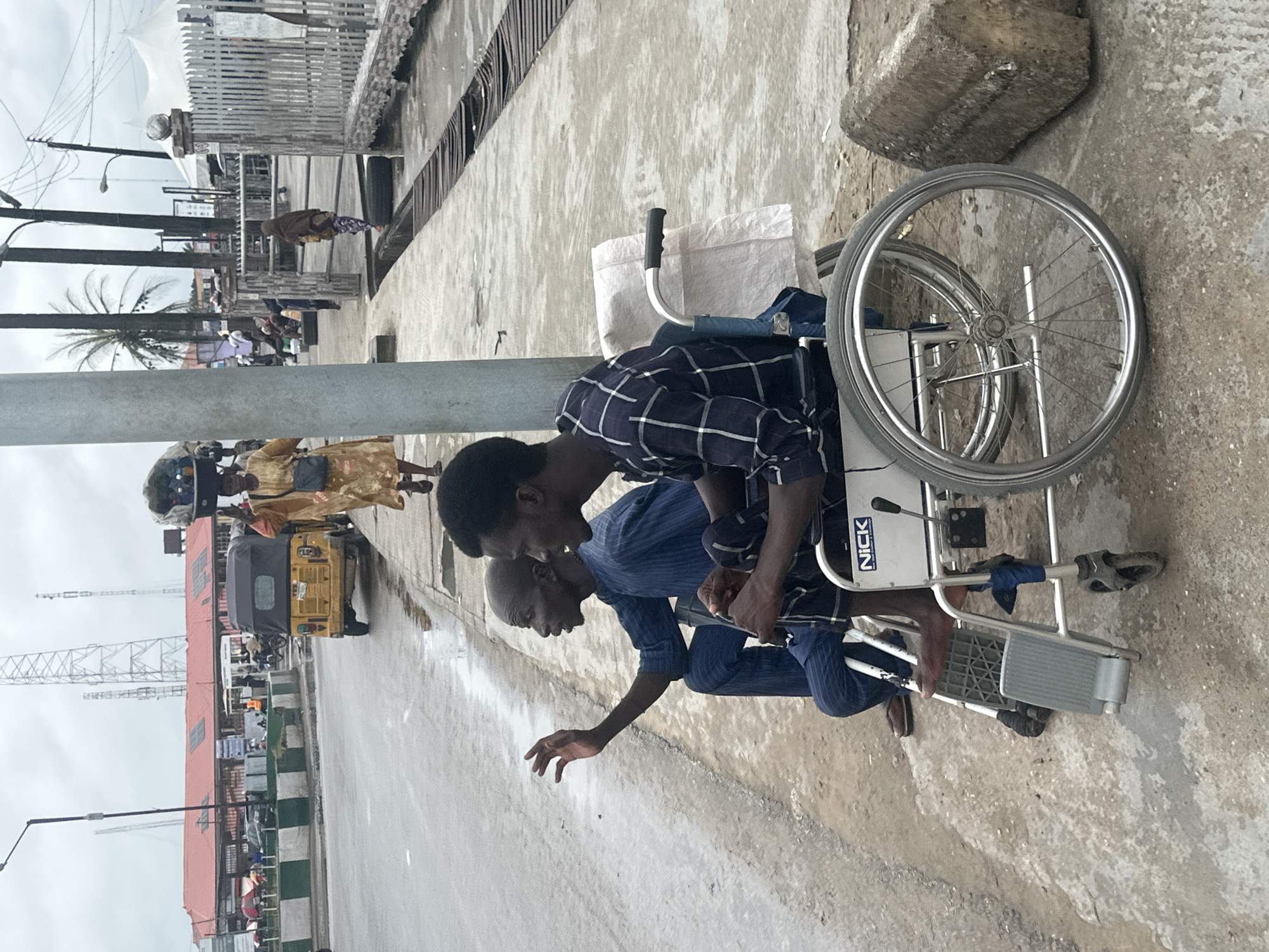 In Badagry, Nigeria, a man in a wheelchair is being assisted by another person. The man in the wheelchair appears to be bald and is wearing a blue striped shirt and dark pants. He is reaching out with his left hand. The person assisting him is wearing a checkered blue and white shirt and dark pants. They are leaning forward, looking at the man in the wheelchair. The wheelchair itself is silver with large spoked wheels. The scene takes place outdoors on a concrete surface, which appears wet, suggesting it may have rained recently. In the background, there are buildings, a yellow motorized tricycle, and other people. There is also a large white bag on the wheelchair. The word "NICK" is visible on the side of the wheelchair. The time of day appears to be daytime, with overcast skies.