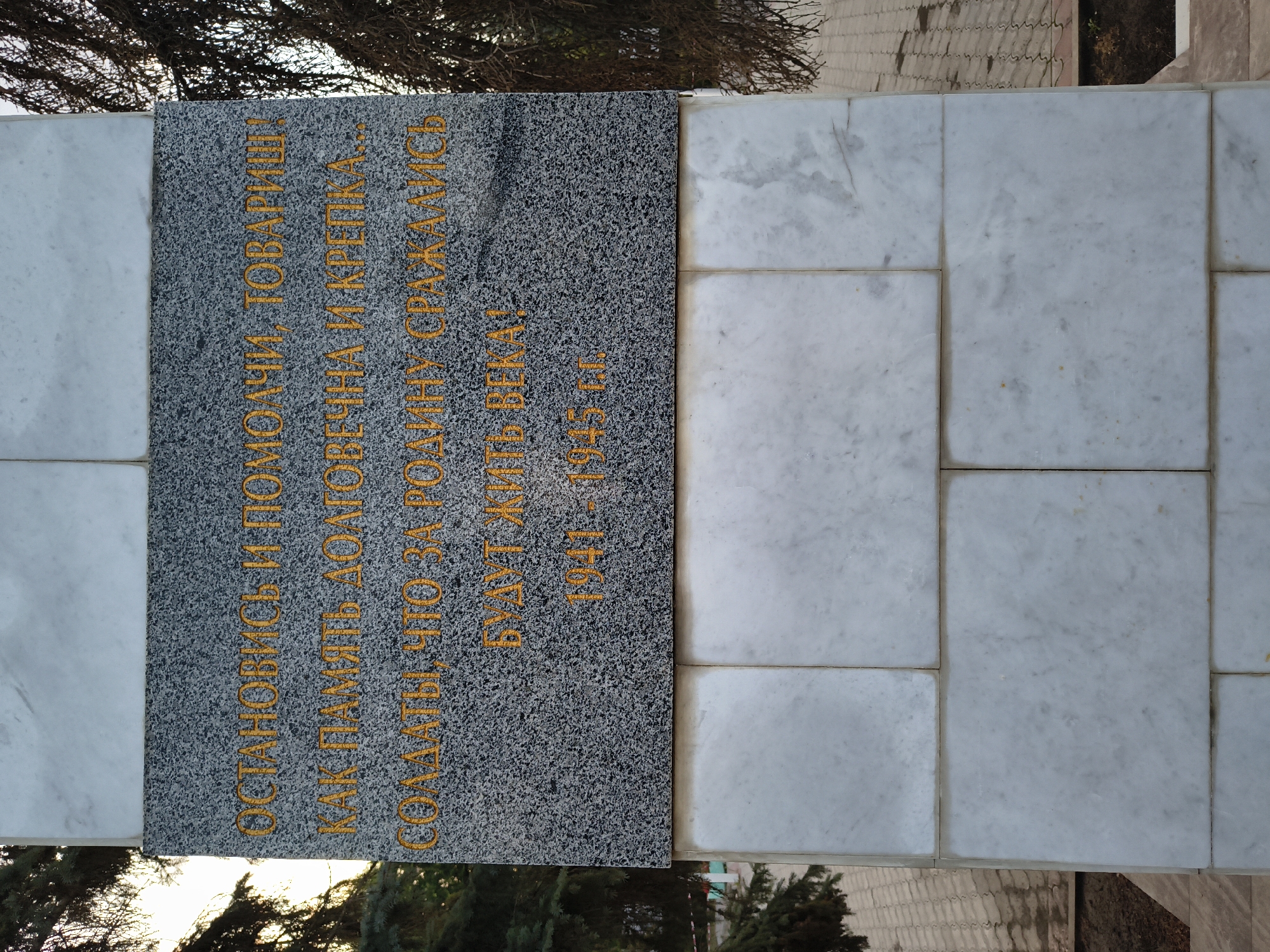 The image shows a monument with an inscription in Russian. The inscription reads: "ОСТАНОВИСЬ И ПОМОЛЧИ, ТОВАРИЩ! КАК ПАМЯТЬ ДОЛГОВЕЧНА И КРЕПКА... СОЛДАТЫ, ЧТО ЗА РОДИНУ СРАЖАЛИСЬ БУДУТ ЖИТЬ ВЕКА! 1941 - 1945 гг." This translates to: "STOP AND BE SILENT, COMRADE! AS MEMORY IS EVERLASTING AND STRONG... SOLDIERS WHO FOUGHT FOR THE MOTHERLAND WILL LIVE FOREVER! 1941 - 1945." The monument appears to be made of granite and marble. The inscription is in gold lettering. The background shows trees and what appears to be a paved area. The monument is located in Mikhaylovka, Russia.