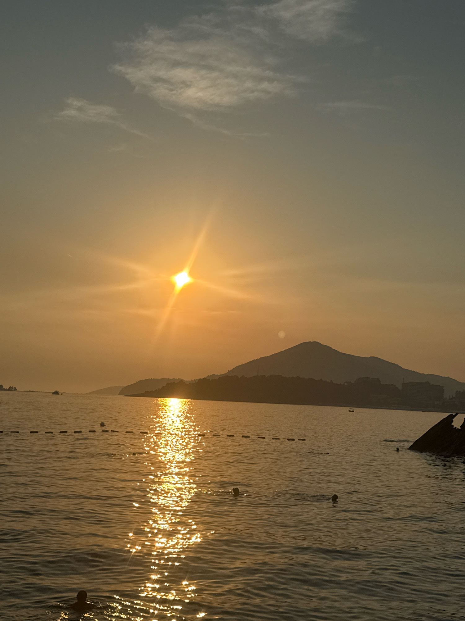 This is a vertical, medium-shot photograph of the sea and sky at sunset, likely in Rafailovići, Montenegro. The sun is low on the horizon, casting a golden glow across the scene. The sun is positioned in the upper left quadrant of the image, emitting bright rays that create a starburst effect. The sea is calm, with gentle ripples reflecting the sunlight, creating a shimmering path that leads towards the foreground.
In the middle ground, a line of buoys with white markers floats on the water, indicating a swimming area. Several individuals are visible in the water, some appearing to be swimming. In the background, a silhouetted mountain range with scattered buildings and a tower on its peak stretches across the horizon. The sky transitions from a warm orange near the sun to a cooler gray at the top of the frame, with wispy clouds scattered throughout. To the right of the frame, a portion of a dark, possibly submerged, object, such as a shipwreck or pier, juts out of the water. A small boat is visible in the distance on the left side of the frame.