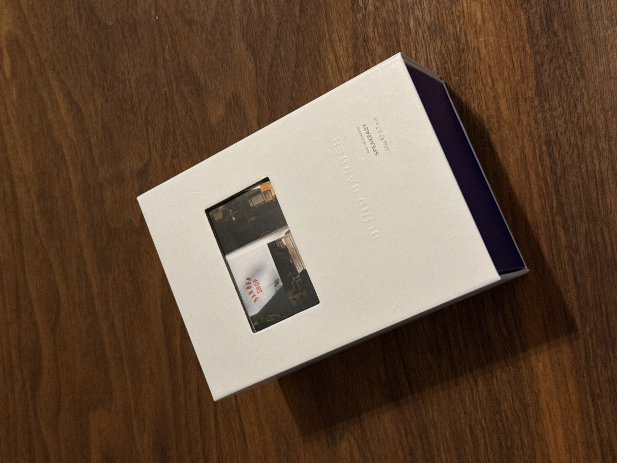 A medium-angle, eye-level shot reveals a white box of "Blind Barber Speakeasy" eau de parfum, set against a dark wooden surface. The box is angled, showcasing a square cutout that reveals a portion of the perfume bottle inside. The box's edge reveals a dark blue interior. The location in Montreal, Canada is not evident from the visible contents.