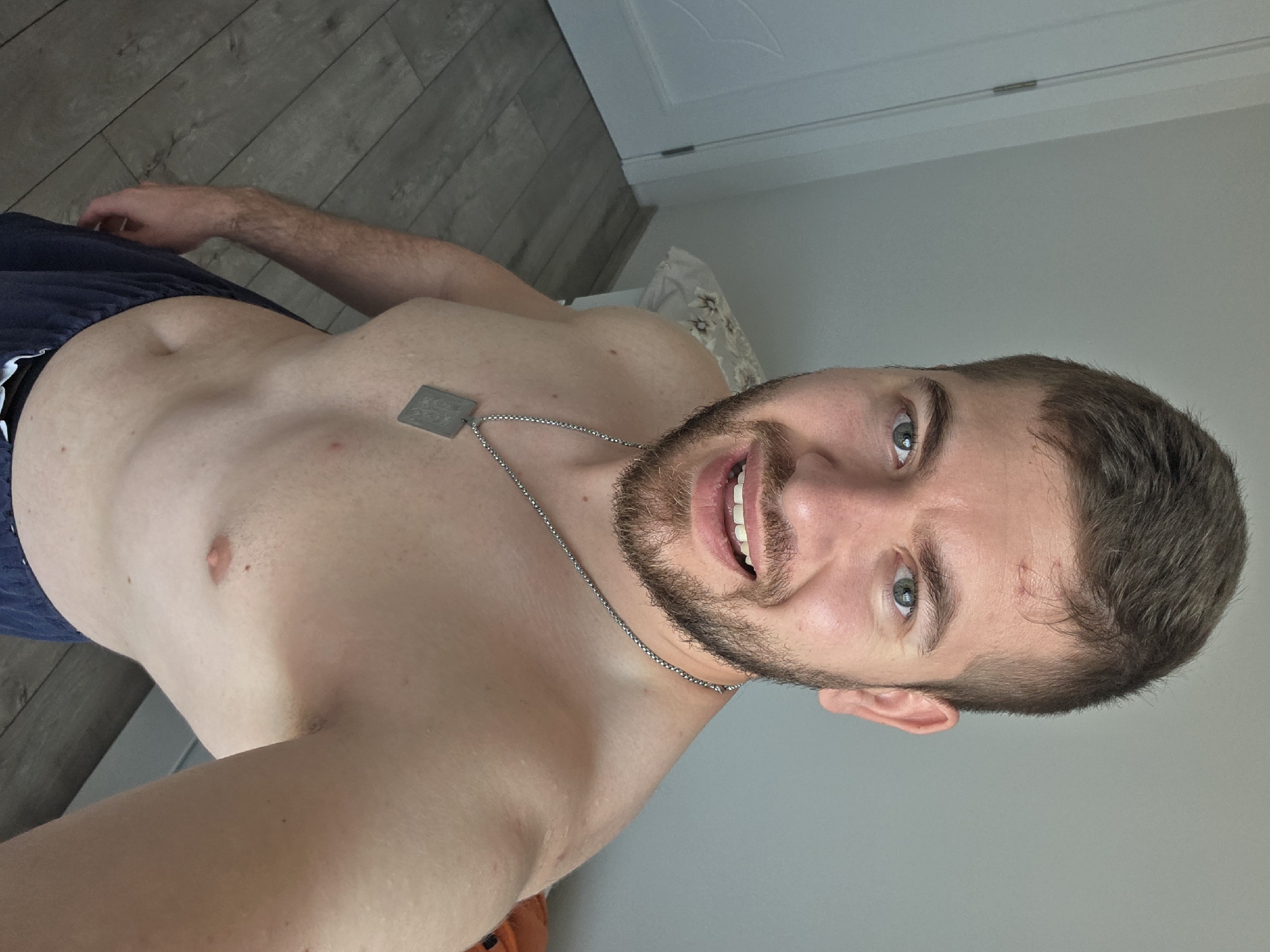 A medium-shot portrait of a man, shirtless, with a dark beard and short brown hair. He is wearing a silver chain with a rectangular dog tag. The man has light skin and green eyes. There is a small cut on his forehead. He is smiling, and his teeth are visible. The background includes a light gray wall, a white door with a decorative panel, and a light wood-patterned floor. The location is unidentified, with no evidence to support its identification as Yagodovo, Bulgaria.