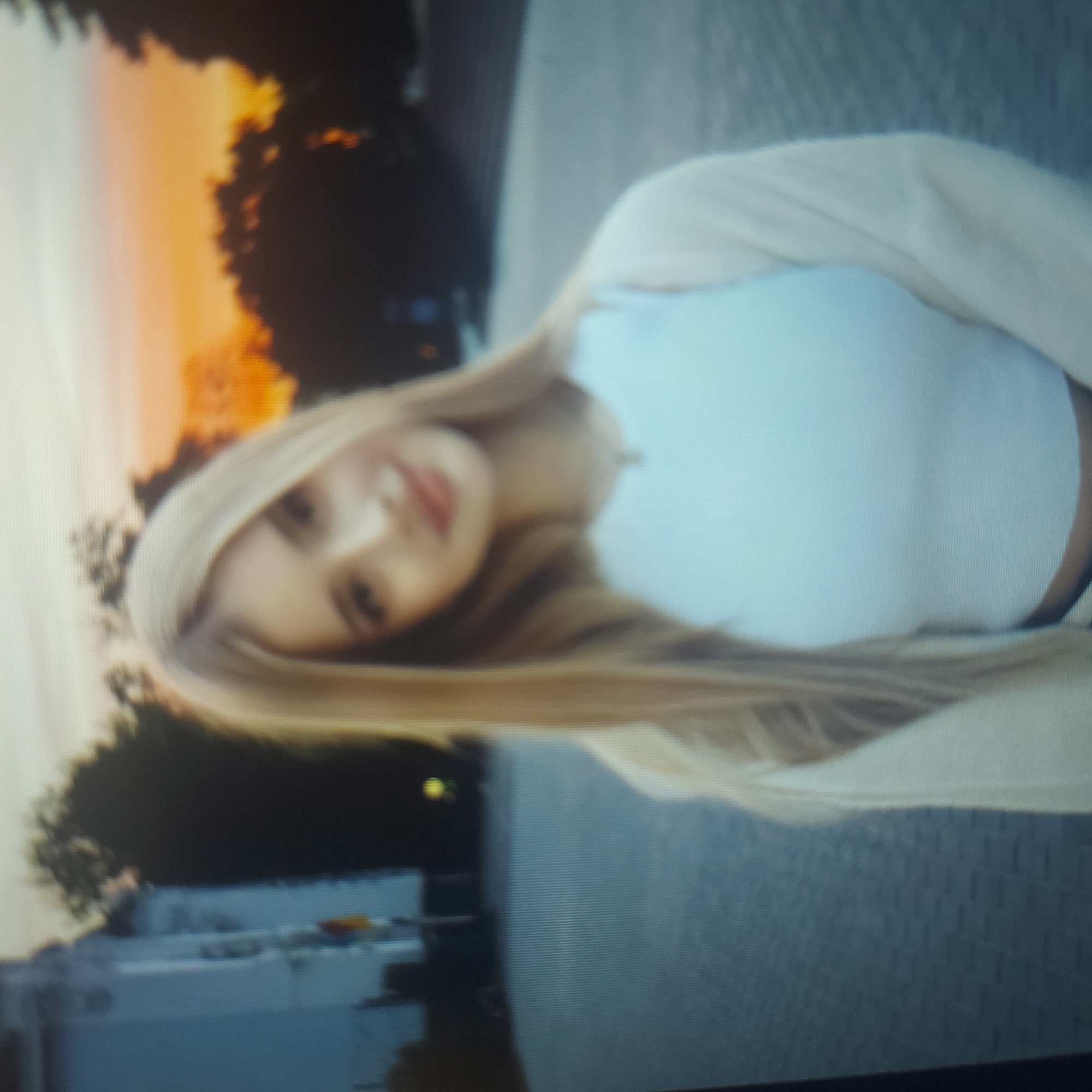 A blurry, pixelated image, appearing to be a photograph of a screen, shows a young woman outdoors. The image is rotated 90 degrees counter-clockwise, with the woman's head positioned on the right side of the frame.
The central subject is a young woman with long, straight, light blonde hair. She is looking towards the viewer with a gentle expression and a slight, closed-lip smile. Her head is tilted slightly. She is wearing a light blue, possibly ribbed, fitted top and a light, likely cream or beige, open cardigan.
The background features silhouetted trees against a warm, gradient sky, transitioning from light orange and yellow hues near the horizon to a lighter, possibly hazy, sky further up. This suggests the time of day is either sunrise or sunset (golden hour). Below the trees, a light-colored building with some darker details is visible. The foreground and mid-ground consist of a dark, textured surface, resembling asphalt or concrete pavement.
The woman is posing for the camera. Her visible emotions are calm and pleasant. There is no visible text in the image. The overall quality suggests it might be a screen capture or a photo taken of a digital display, as evidenced by the visible pixel grid and slight blurriness.