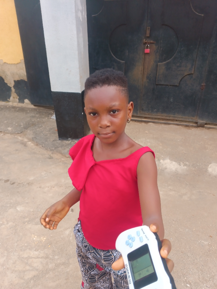 A young girl stands outdoors in Itu, Nigeria, facing the camera with a neutral expression. She has dark skin, short dark hair with a faded side part, and wears a small hoop earring in her left ear. Her attire consists of a red sleeveless top with a decorative ruffle on the left shoulder/chest and patterned trousers featuring black, white, and red abstract designs. Her right arm is extended towards the viewer, holding a white handheld electronic game device. The device has a rectangular monochrome screen displaying pixelated graphics, four blue directional buttons on the left, and four blue action buttons on the right, with "GAME TOY II" visible on its casing. The ground beneath her is a light-colored, textured surface. In the background, to the left, is a yellow wall partially obscured by a white rectangular column with a black base. To the right, a dark, weathered double-leaf gate with decorative elements is secured by a red padlock in the center.