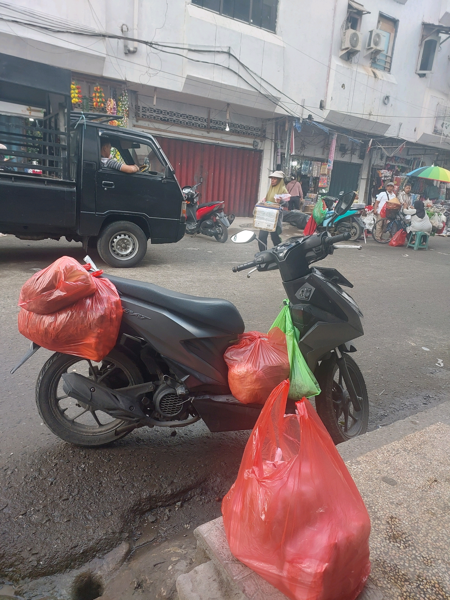 The image depicts a street scene in Ambon, Indonesia, likely a market area, characterized by its bustling and somewhat chaotic atmosphere. In the foreground, a dark-colored scooter is parked, laden with several large, bright red plastic bags, suggesting a recent purchase or delivery. Two green plastic bags are also attached to the scooter's handlebars, possibly containing smaller items.
Behind the scooter, a black pickup truck is visible. The driver, a man wearing a striped t-shirt, is seated behind the wheel, looking forward. The truck's bed is open and appears to be transporting items, though they are not clearly discernible. In the background, the architecture suggests a commercial area with shops and stalls lining the street. Several individuals are present, some walking, others engaged in what appears to be market activities, including a person holding a box and others near a stall with an umbrella. The ground is paved with asphalt and shows signs of wear and tear, with some muddy patches and unevenness. The overall lighting indicates a daytime scene, with clear shadows cast by the objects.