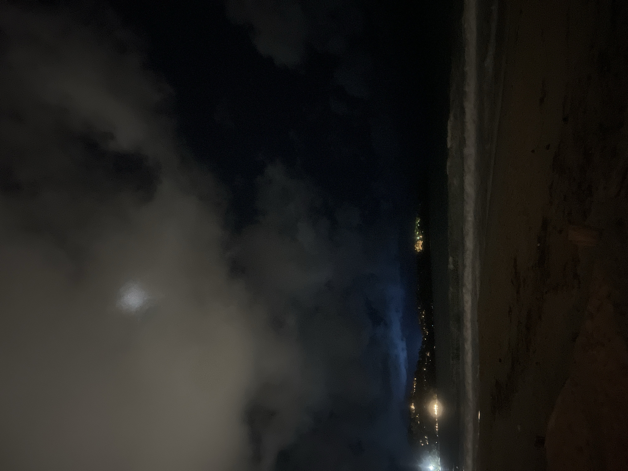 The image shows a night scene at a beach in Unknown, Unknown. On the right, there is a beach with the waves on the edge. On the left, there are clouds, and there appears to be a moon behind them. There are lights that could be from a city in the distance along the beach.