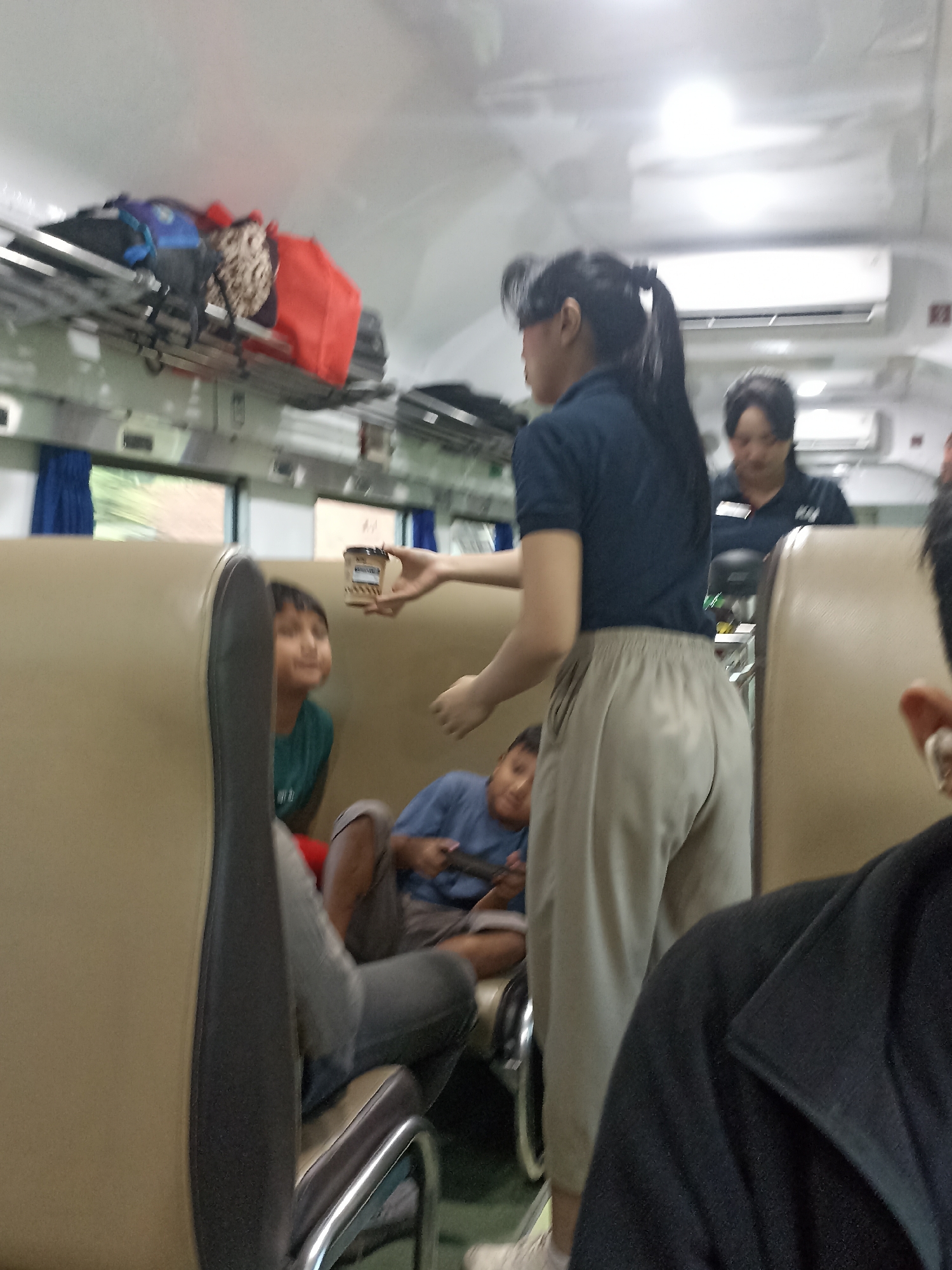 In a train carriage in Kaligondo, Indonesia, a woman in a dark blue polo shirt and light-colored pants offers a small brown container to a young boy in a green shirt who is seated. Another young boy in a blue shirt is seated nearby, looking towards the woman. Luggage is stored on overhead racks. The train carriage appears to be in motion, indicated by the blurred background through the windows.