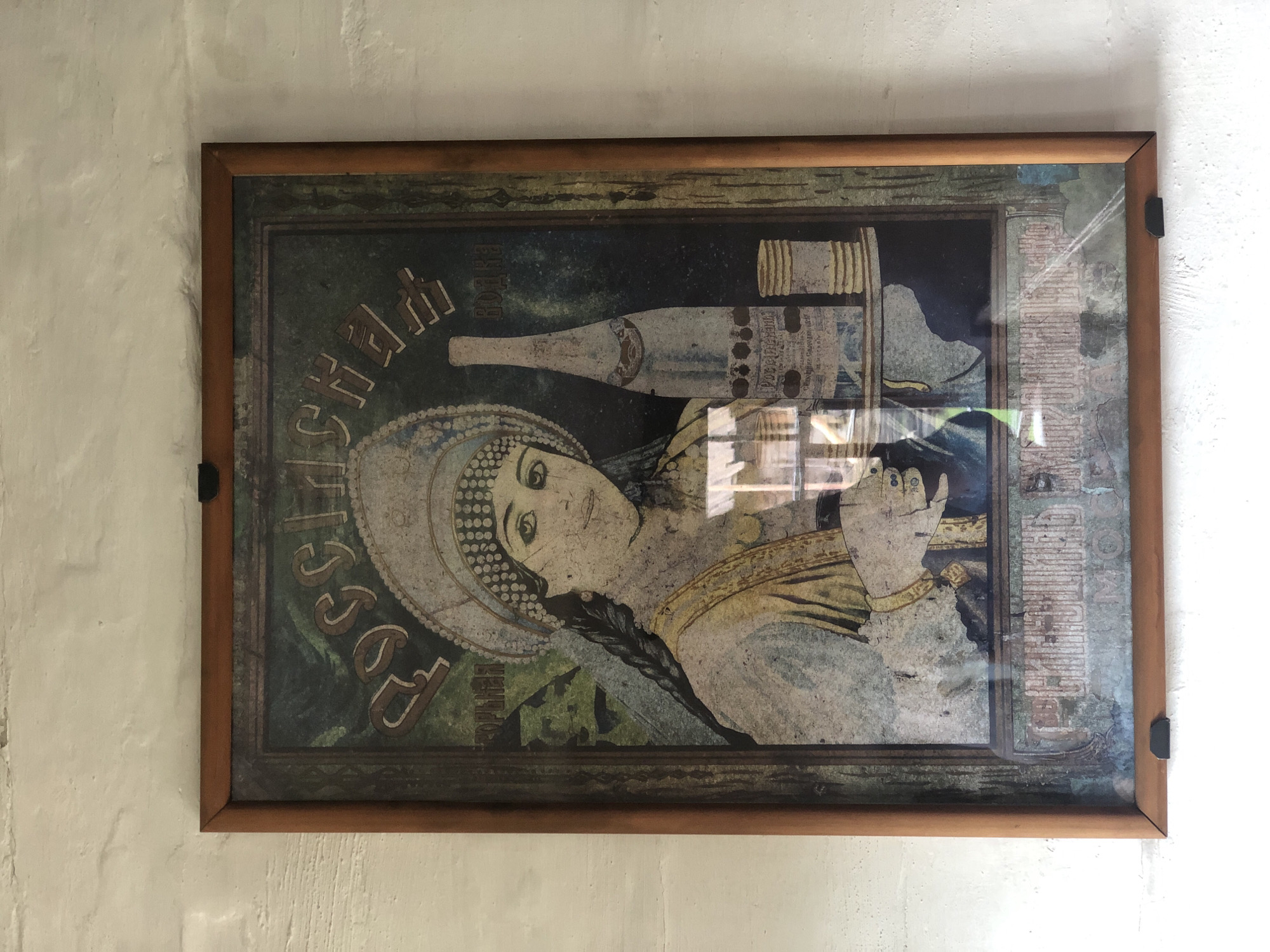 An antique framed poster depicts a woman in traditional Russian attire, possibly a kokoshnik, holding a tray. On the tray are a bottle of what appears to be vodka and several stacked coins or discs. The background is a dark, textured green, with Cyrillic lettering at the top. The poster is mounted on a white brick wall. The scene is the interior of a building in the city of Shushenskoye, Russia.