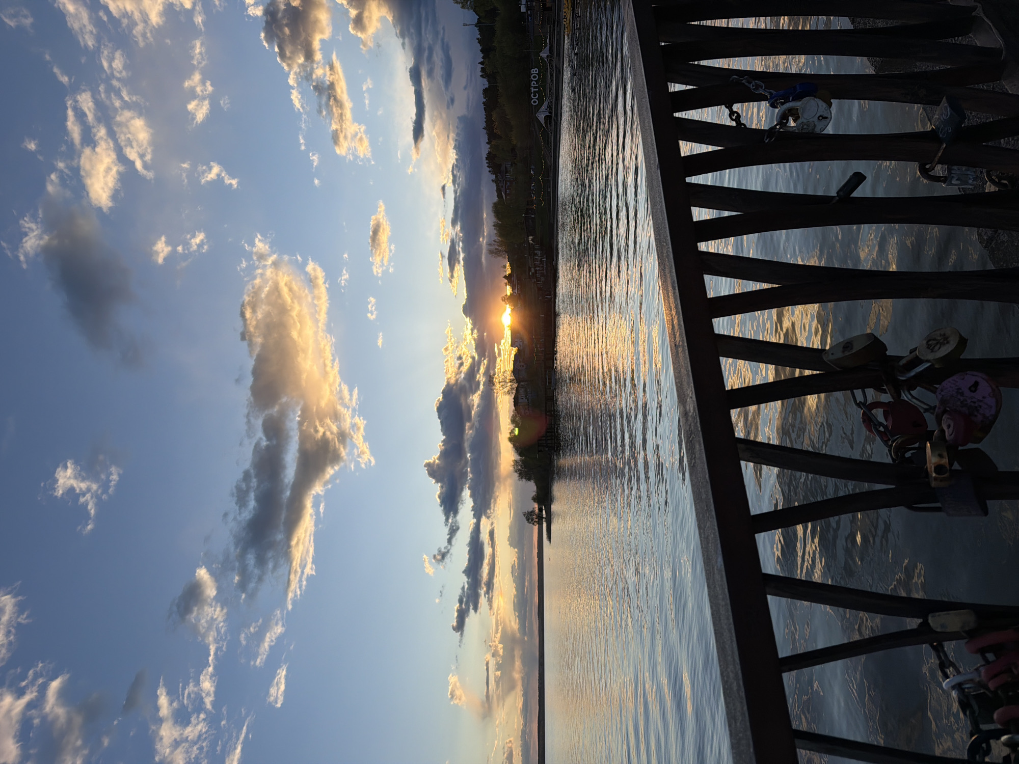 The image depicts a sunset over a body of water from what appears to be a bridge in Votkinsk, Russia. The sun is setting behind a line of trees on the horizon, casting a golden light across the water. The sky is filled with scattered clouds, some illuminated by the setting sun. The railing of the bridge is visible in the foreground, and it has several padlocks attached to it.