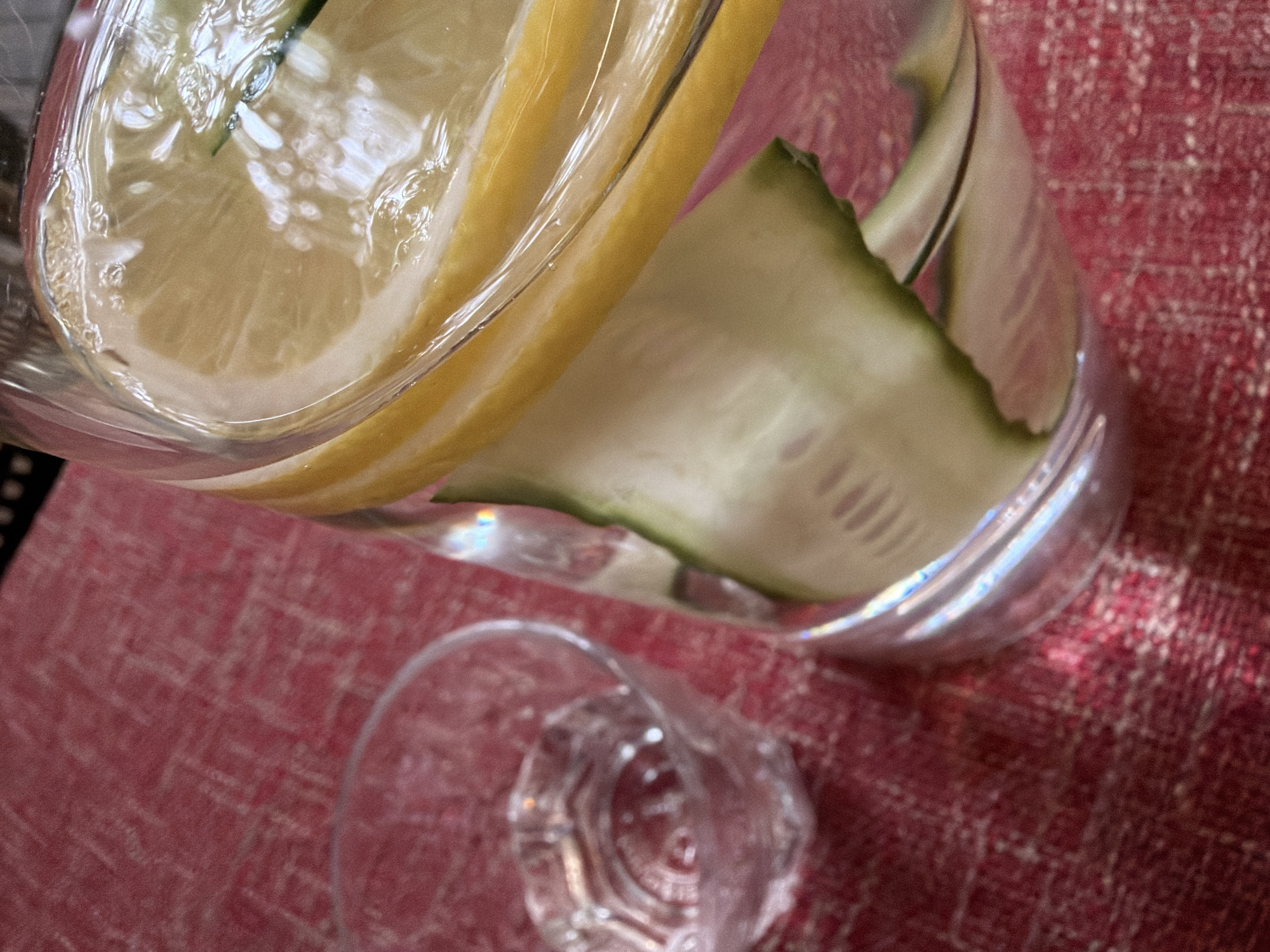 An overhead shot shows a clear glass pitcher filled with water, lemon slices, and cucumber slices. To the left of the pitcher is a small, empty clear glass. Both are situated on a textured red tablecloth. The pitcher is positioned on the right side of the frame, taking up a significant portion of the image. The water inside the pitcher appears to have small bubbles and reflections. Slices of yellow lemon are layered near the top, followed by slices of green cucumber. The tablecloth has a woven pattern and is lit from above, casting shadows and highlights. There are no people visible in the image. The scene is a close-up view of a refreshing drink preparation or presentation.