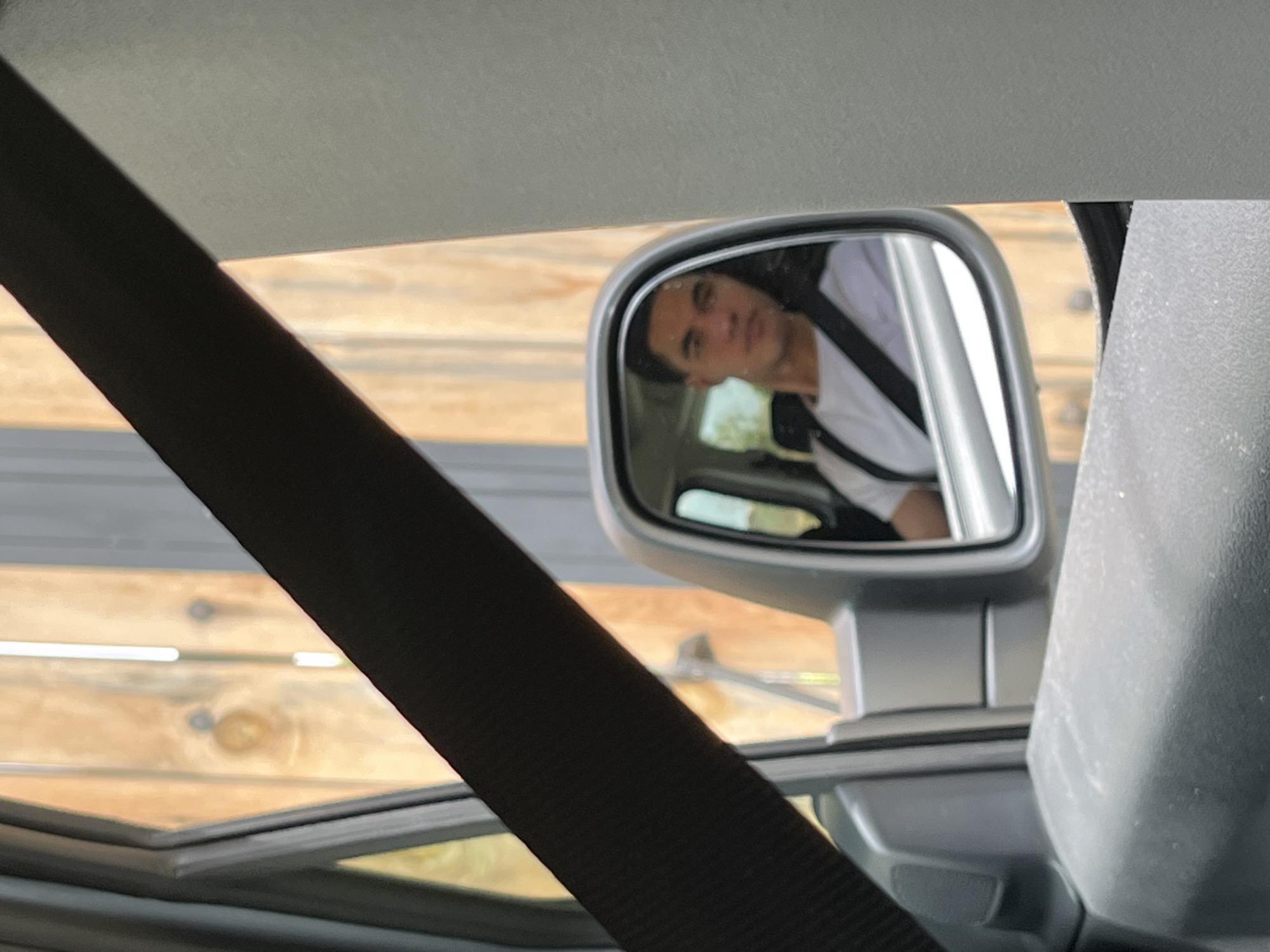 A man is visible in the passenger side mirror of a vehicle. He wears a white t-shirt and a black seatbelt. The background visible through the car windows appears to be wooden boards with nails or fasteners. The interior of the car is visible, including the seatbelt and car frame. The location is not discernible from the image.