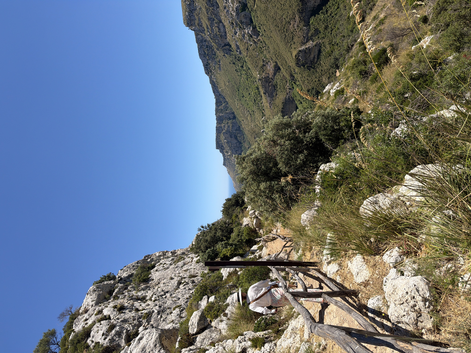 A woman wearing a wide-brimmed hat and a patterned skirt walks along a rocky path with a wooden railing. She is seen from behind, her right shoulder facing the camera. The path is on a hillside with sparse vegetation, including dry grasses and green shrubs. To the left, a steep, rocky cliff rises up, dotted with hardy green plants. To the right, the path descends into a lush, green valley with more rugged, terraced hillsides stretching into the distance under a clear blue sky. The scene appears to be outdoors in a natural, mountainous environment. This location is the Vendicari Nature Reserve, near Noto, Italy.