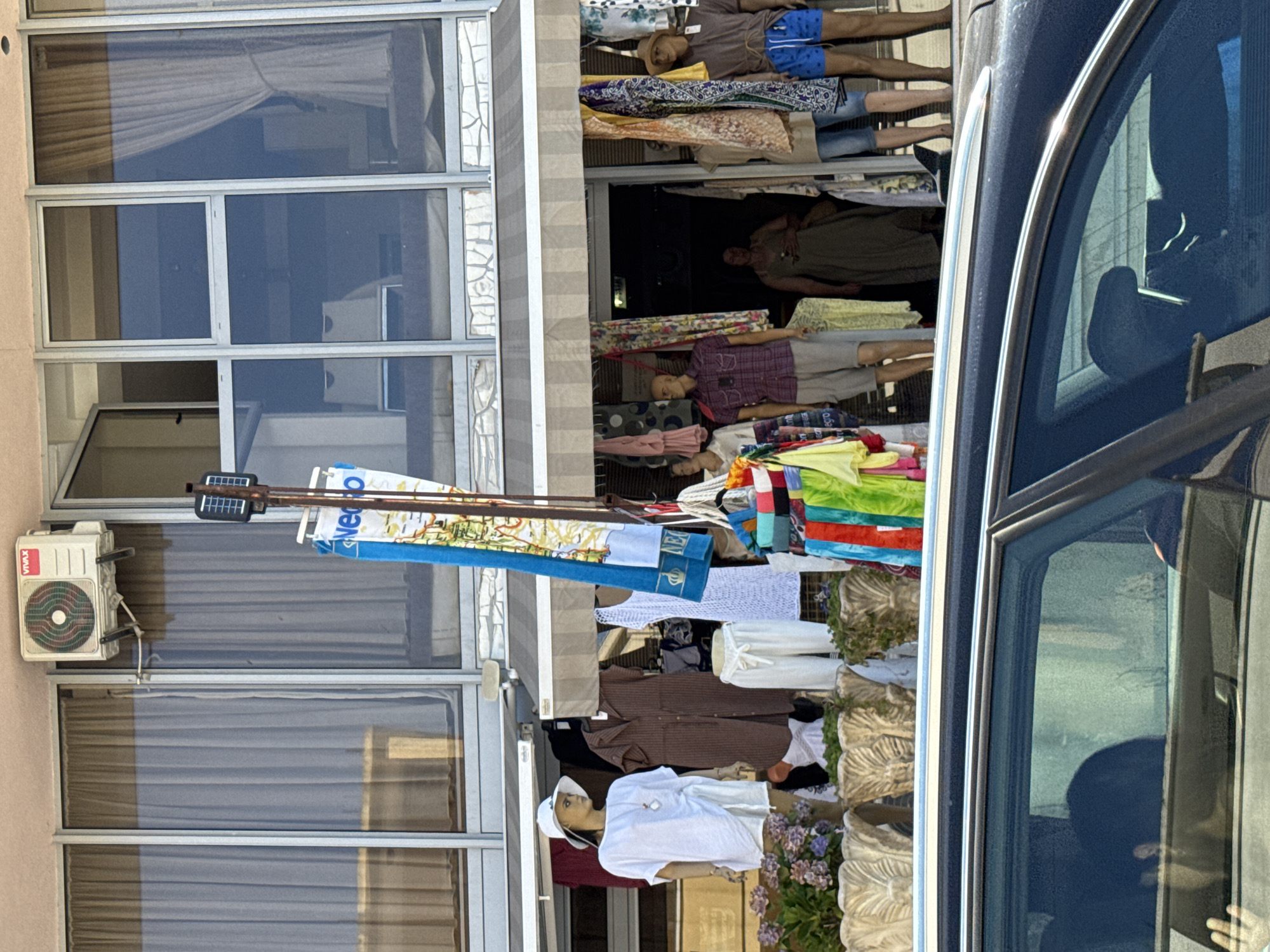 The image shows a storefront in Budva, Montenegro, displaying clothing on mannequins and racks. In the foreground, a car is partially visible, with its roof rack and tinted windows reflecting the scene. The storefront features large glass windows and an awning. Mannequins are dressed in casual summer clothing, including dresses, shorts, and hats. Colorful fabrics and beach towels are also displayed. An air conditioning unit is mounted on the wall above the windows. A woman is visible inside the store, and another person can be seen in the car. The scene appears to be on a sunny day.