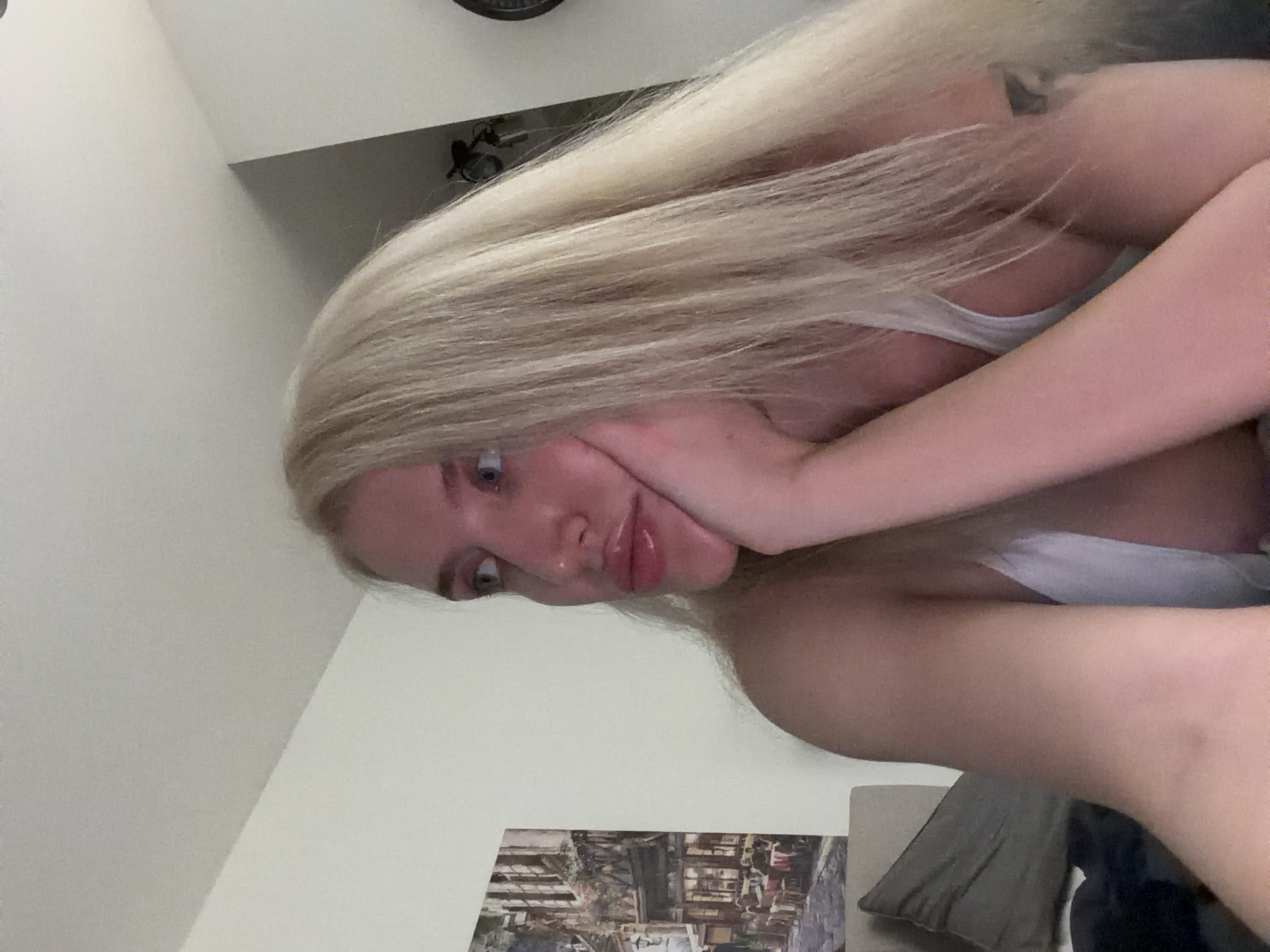 An indoor portrait displays a young adult female, positioned in a three-quarter profile facing left. She possesses long, straight blonde hair and fair skin. Her light blue eyes are directed upwards and to her left. Her left hand is resting against her chin and jawline, fingers lightly curled. She is wearing a white tank top. A dark, stylized tattoo is partially visible on her right bicep/shoulder. The background to her left features a wall-mounted framed artwork depicting an old-world European street scene with buildings and outdoor seating. Below the artwork, a dark grey pillow is partially visible. The background to her right consists of a light-colored wall, with a partial view of a dark, circular object, possibly a wall clock, in the upper right corner. The specific location within the city of Belgrade, Serbia, is not visually discernible from the details present in the image.