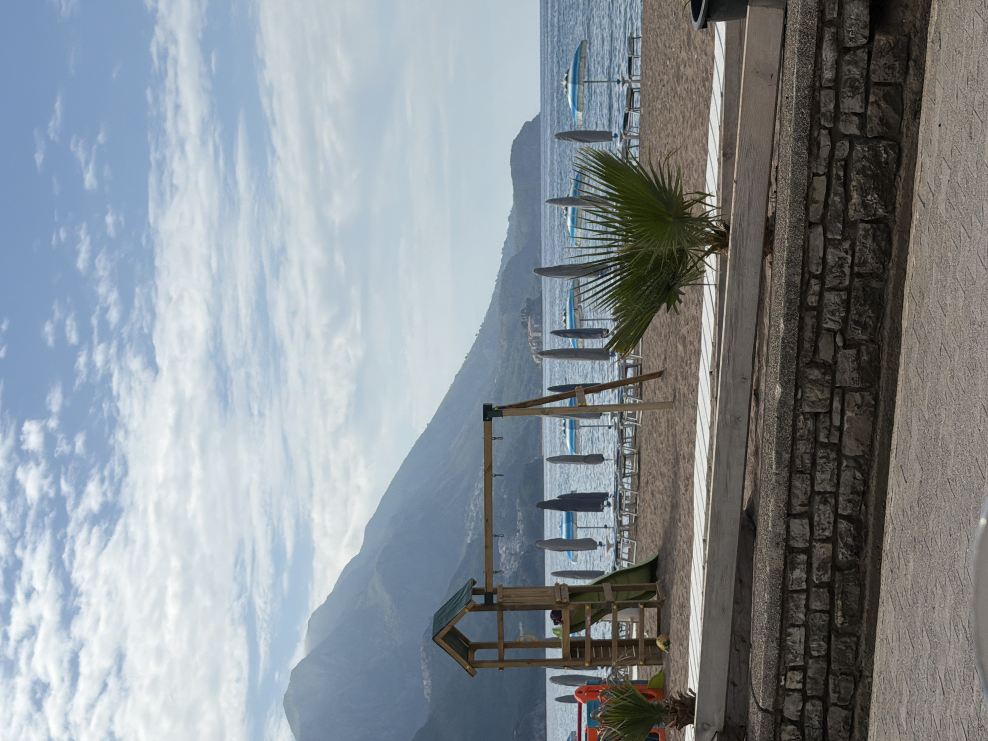 The image shows a scene at a beach near the city of Boreti, Montenegro. The sky is light blue and filled with patchy white clouds. Adjacent to the beach is a stone wall. The beach has rows of lounge chairs covered by umbrellas. In the middle ground is a set of wooden playground equipment with a slide and swings. There is a lush green mountain in the background. A palm tree is at the edge of the scene near the wall. The beach leads into the sea and the ocean.