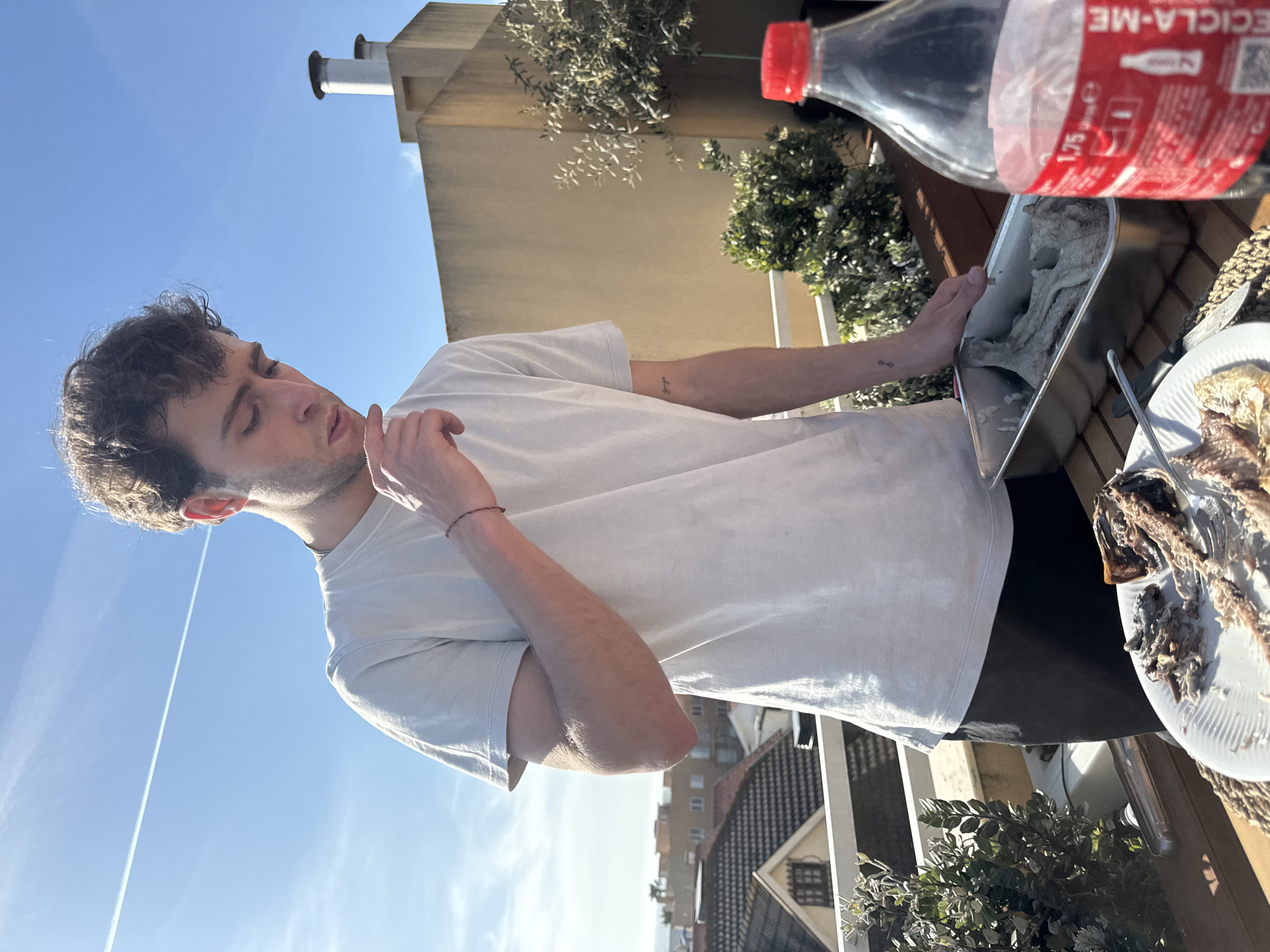 A man with short brown hair stands on a balcony, holding a metal container. The container appears to hold food and is situated near a plate of consumed food remains. A large bottle of Coca-Cola is present in the frame.
The environment shows a cityscape, indicating an urban residential area. In the background, one can see buildings and greenery typical of Vila Nova de Gaia, Portugal. A blue sky is above.
