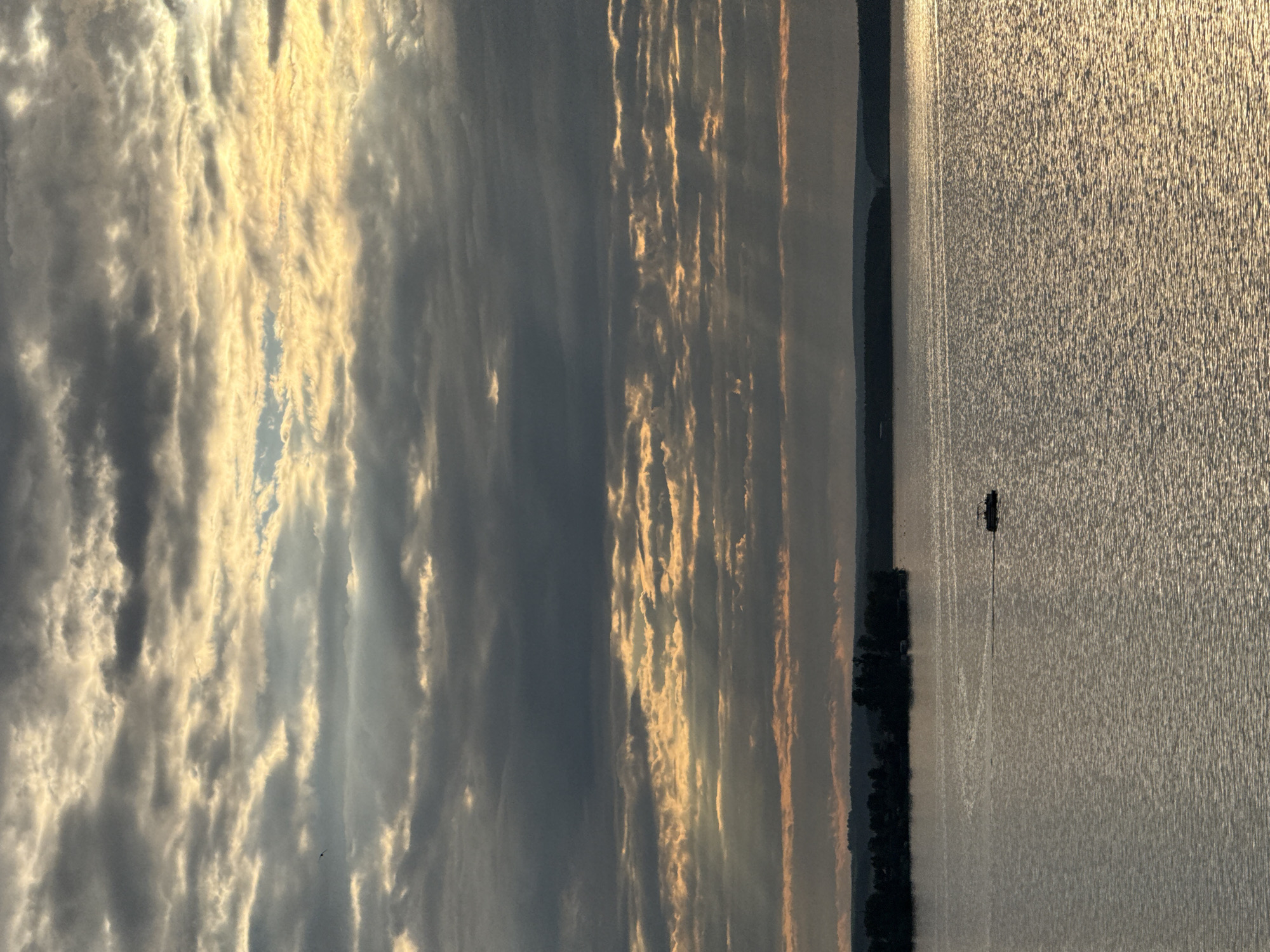 This image depicts a body of water with a pontoon boat in the distance, under a sky filled with layered clouds illuminated by sunlight. The water is rippled, reflecting the light and sky. In the background, a tree-lined shore is visible, with a dark silhouette of hills beyond. A small, dark object, possibly a bird, is visible in the upper left quadrant of the sky. The lighting suggests either sunrise or sunset. This scene is located on the Votkinsk Reservoir in Votkinsk, Russia. There are no people visible in the immediate vicinity of the boat.