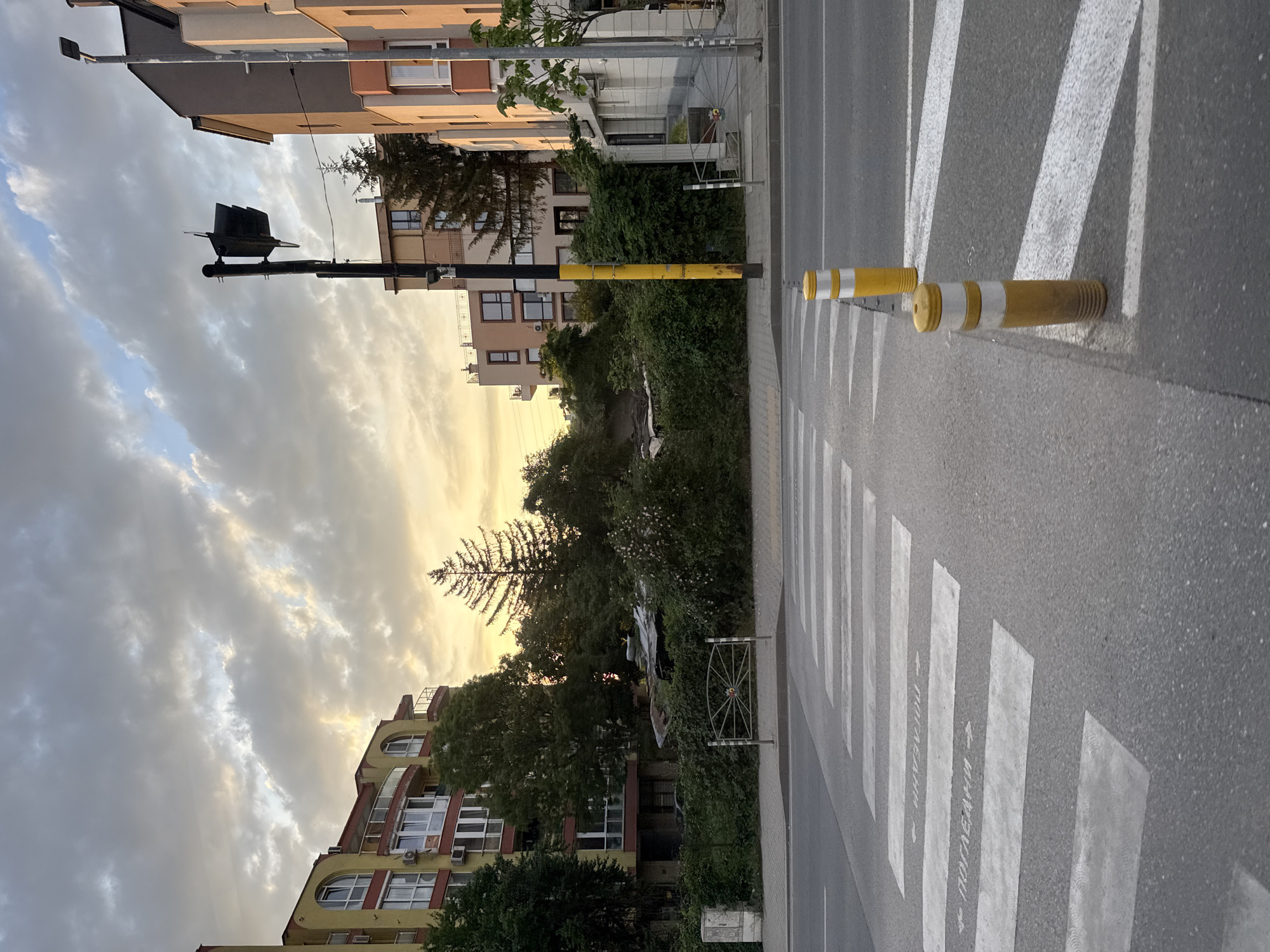 The image depicts a street scene in Sofia, Bulgaria. A pedestrian crosswalk stretches across the asphalt road, marked with alternating white stripes. A pair of short, yellow and white bollards are installed on the right side of the crosswalk, likely to deter vehicles from parking or encroaching. The text "ПОГЛЕДНИ" with arrows is written on the white stripes, which translate to "Look." Buildings line the left side of the street, including residential structures with windows and balconies. Green foliage is present, suggesting trees or shrubs. A street light and traffic signal are visible on the left. The sky is partially cloudy, with a hint of orange.
